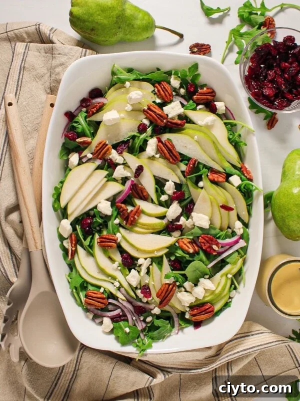 A large white serving dish filled with an arugula and pear salad with toasted pecans, feta cheese, dried cranberries and red onion.