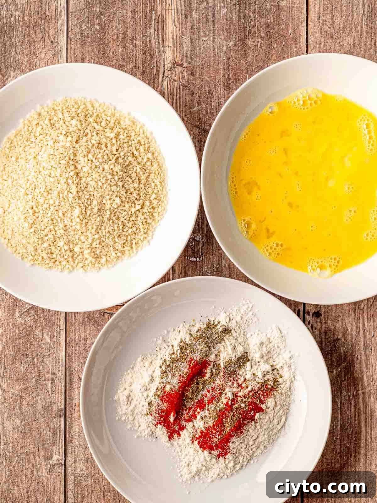 A bowl of breadcrumbs, a bowl of eggs and a bowl of flour with spices on a table.
