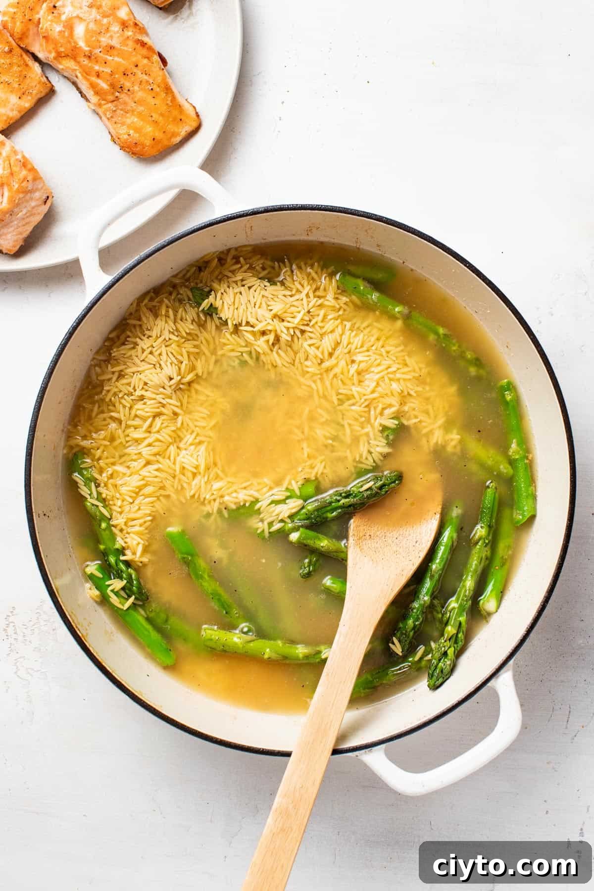 Adding orzo and broth to a pan with fresh asparagus.