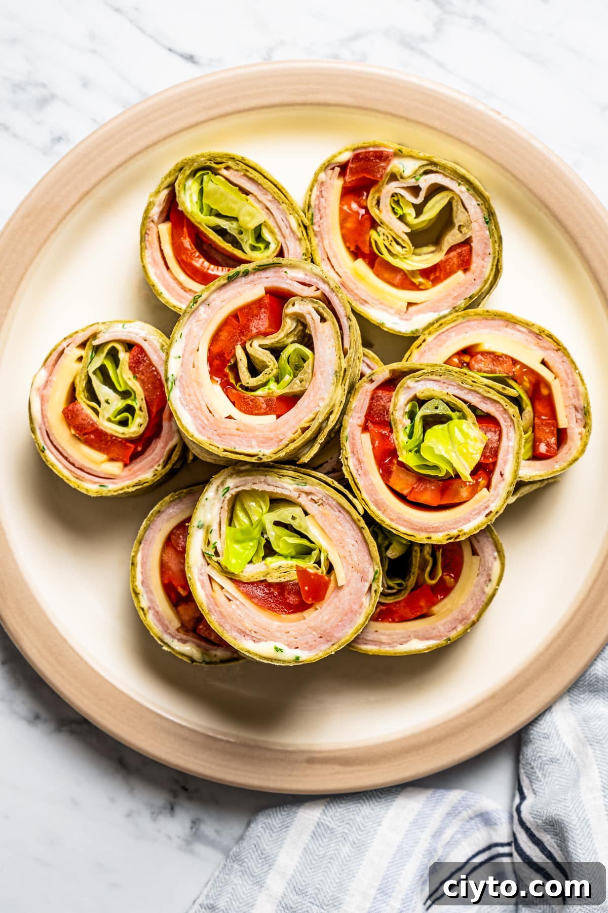 Pinwheel sandwiches are loaded with sliced meats, cheeses, vegetable matter, and dijon-dill mayo and sliced into perfect 2-bite morsels. Overhead image of pinwheel sandwiches arranged on a serving plate.