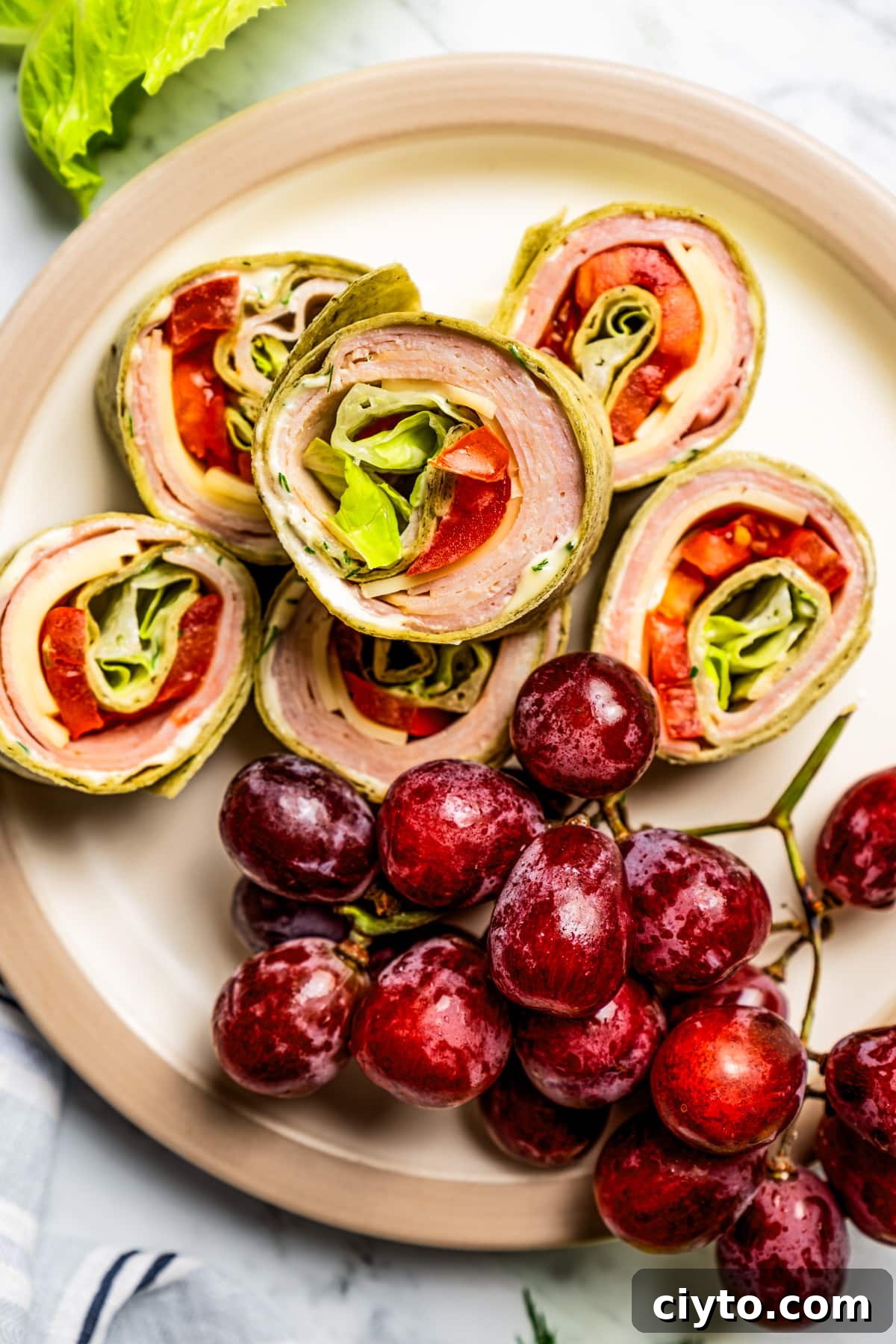 Pinwheel sandwiches paired with fruit for a simple plated lunch. Closeup of pinwheel sandwiches stacked on a plate with a bunch of grapes.