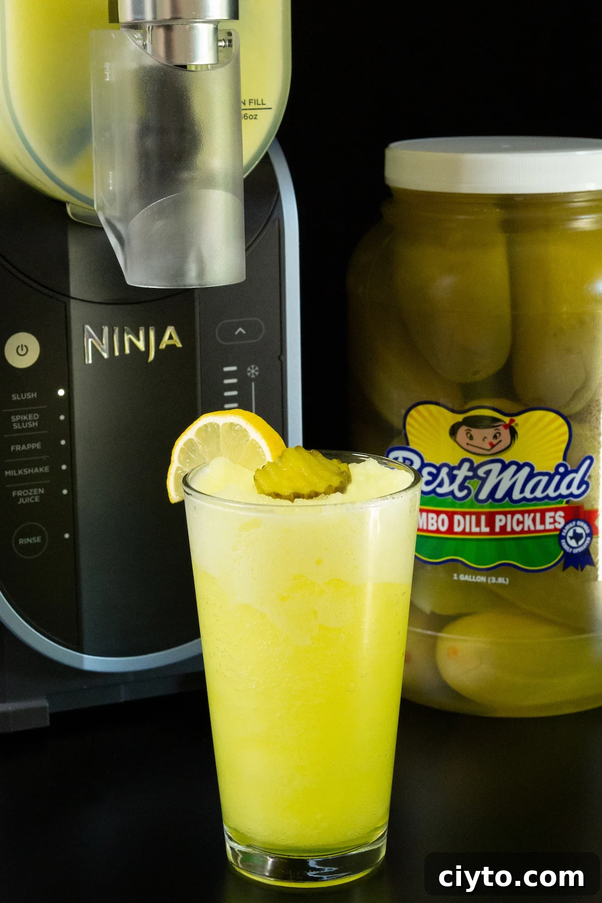 A pint glass filled with a bright yellow pickle slushi sits in front of a large jar of dill pickles and the Ninja Slushi machine.