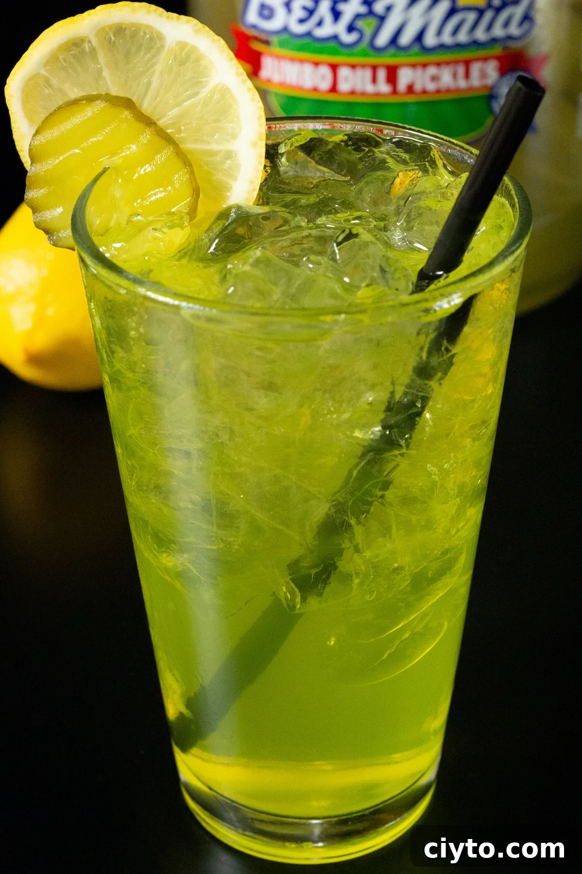 A pint glass filled with clear ice cubes and a bright yellow pickle lemonade drink that's garnished with a pickle chip and a lemon wheel.