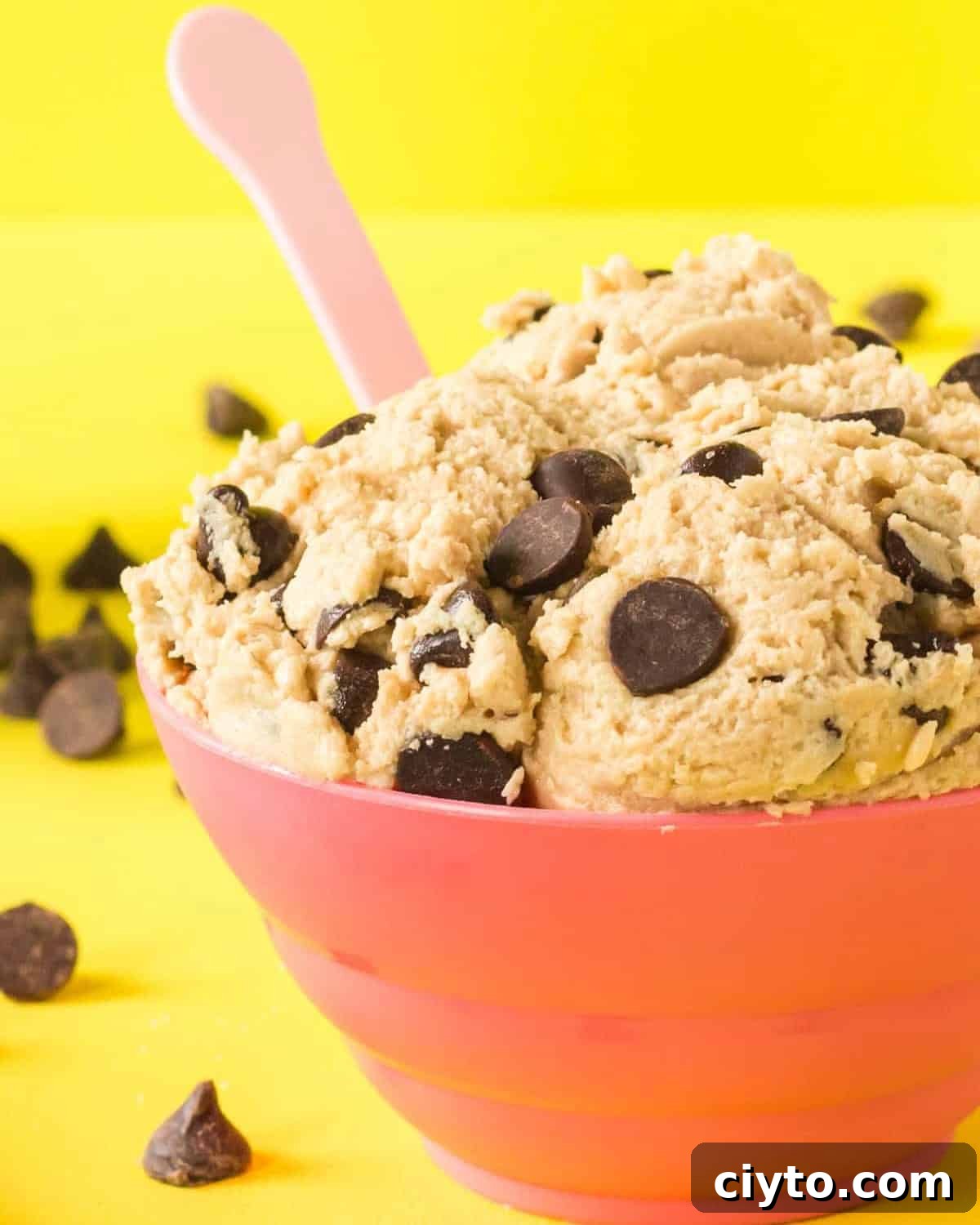 closeup of a bowl of edible chocolate chip cookie dough with a spoon on a yellow background.