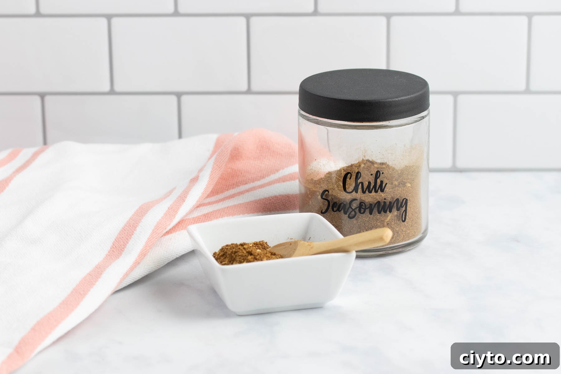 Jar of homemade chili seasoning beside a dish of the seasoning with a small wooden spoon.