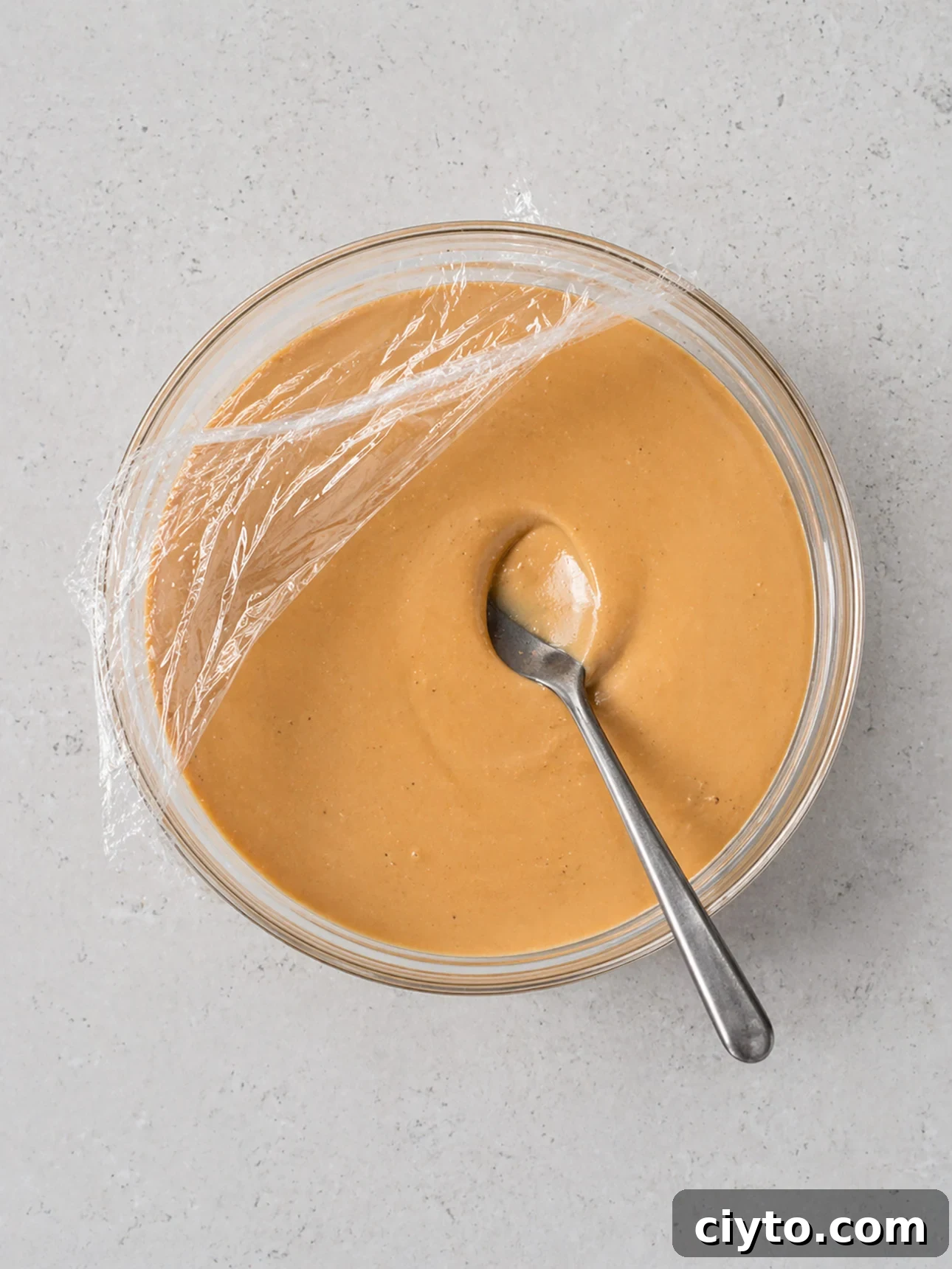 A glass bowl filled with creamy salted caramel gelato, partially covered with plastic wrap, and a metal spoon resting inside on a light gray surface.