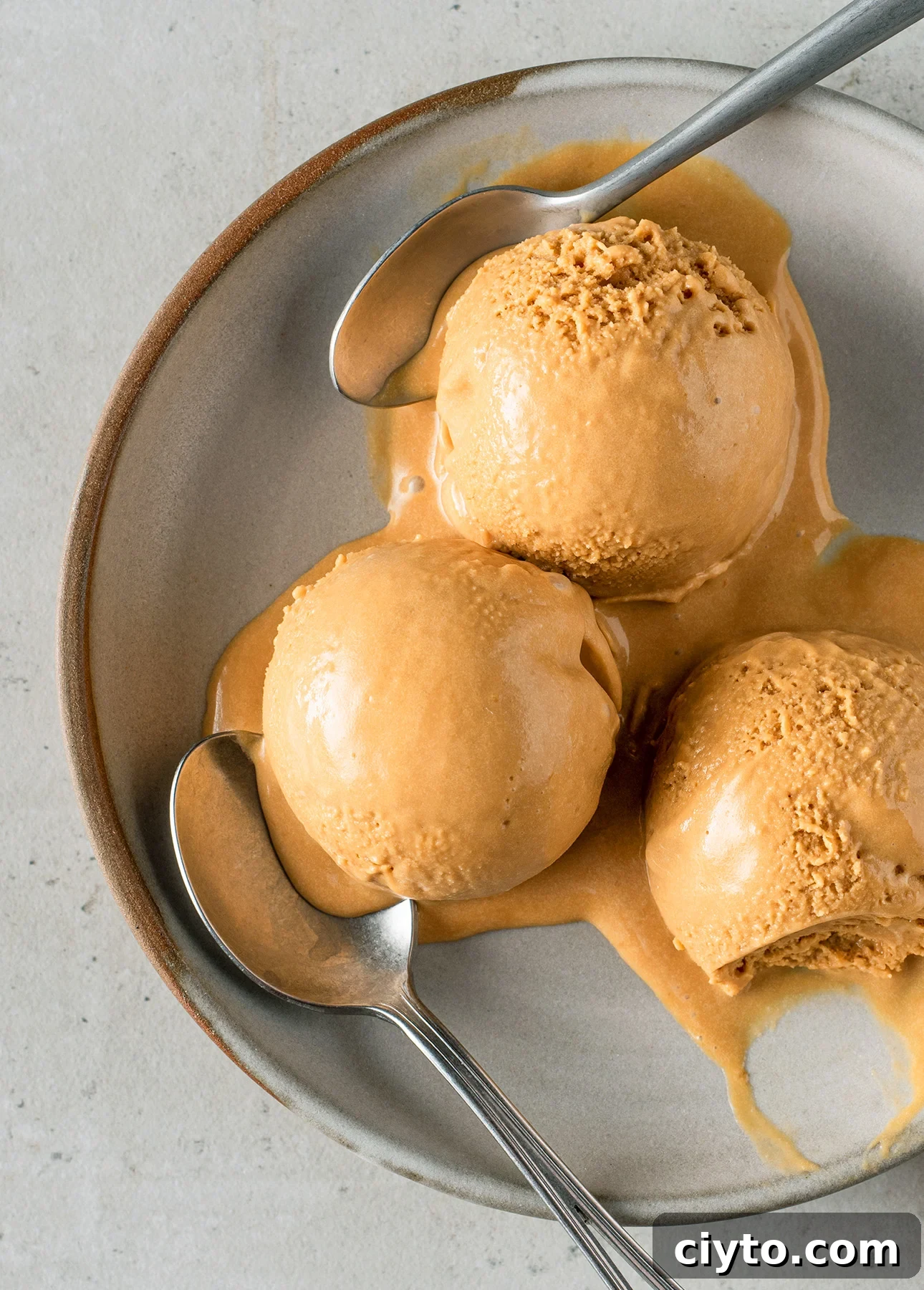 Three scoops of salted caramel gelato are melting on a ceramic plate, with two metal spoons resting beside and partially on the creamy, caramel-colored scoops.