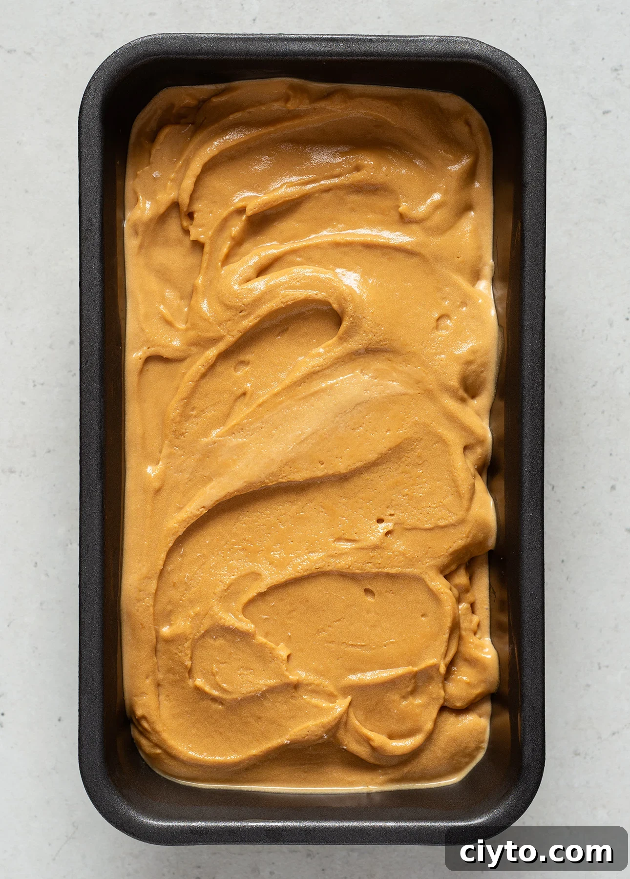 A rectangular metal loaf pan filled with smooth, creamy salted caramel gelato, its rich caramel-colored swirls atop a frozen dessert, set on a light surface.