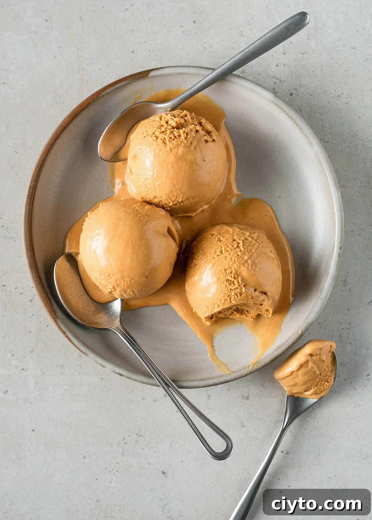 Three scoops of melted salted caramel gelato on a round white plate, with three silver spoons placed around and on the gelato. The gelato is melting, creating rich caramel-colored pools on the plate.