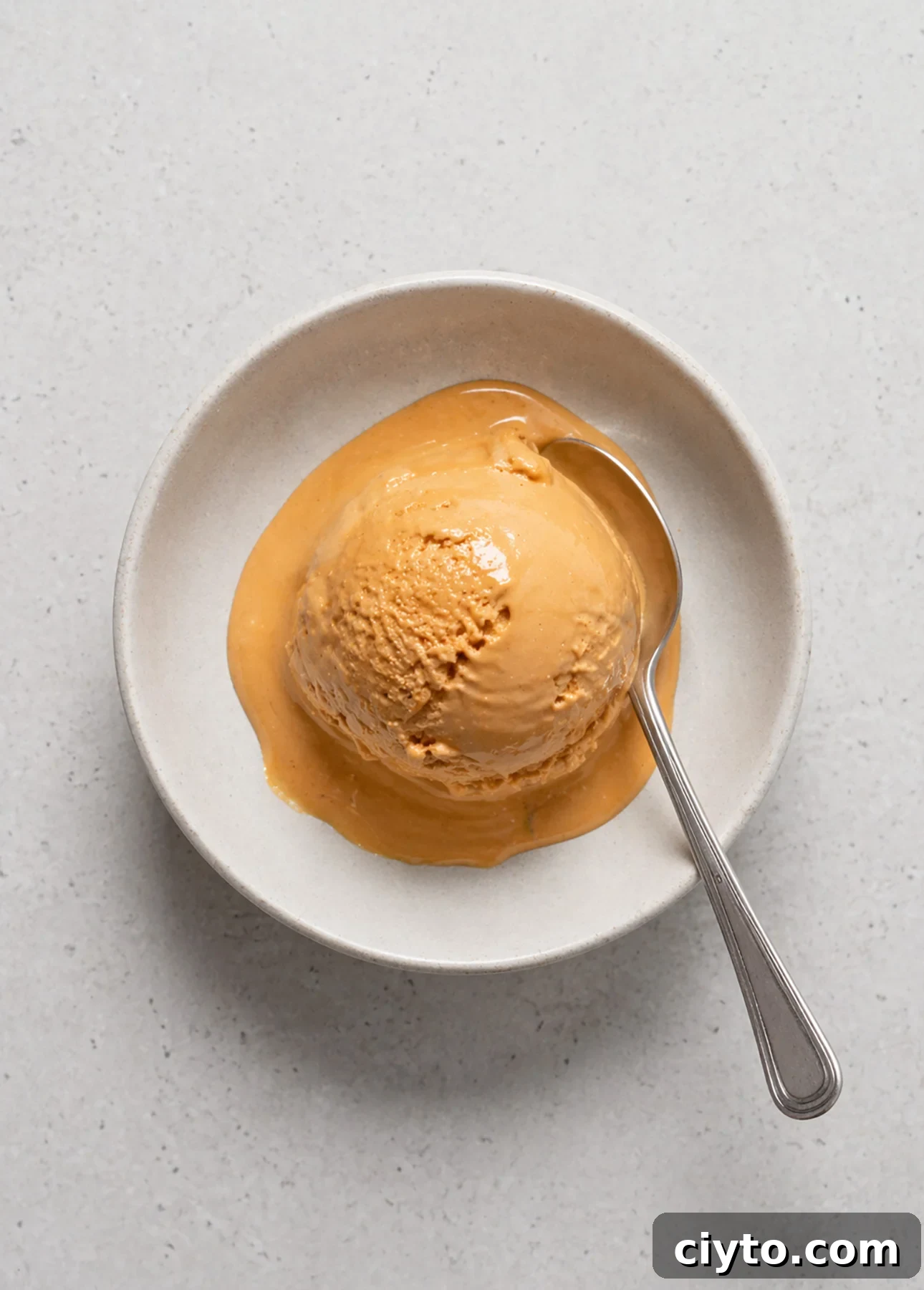 A bowl of salted caramel gelato is melting, with a spoon resting on the side, all set on a light, textured surface.