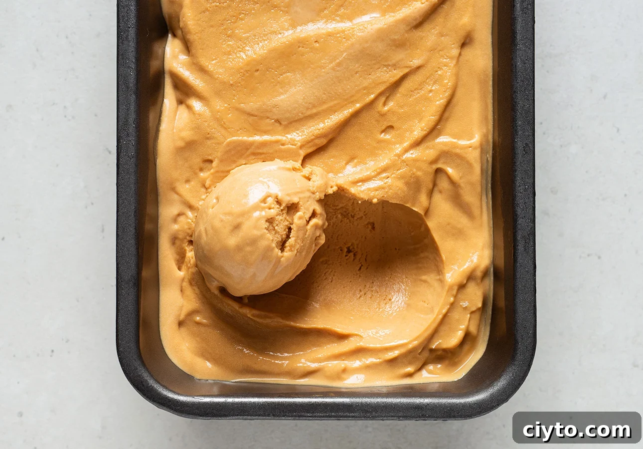 A rectangular metal pan filled with creamy, light brown salted caramel gelato, with one scoop taken out and resting on top of the smooth surface.