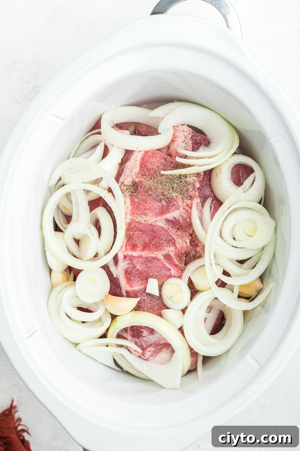 Uncooked ribs in a white slow cooker, topped with onion slices, garlic, and seasonings.