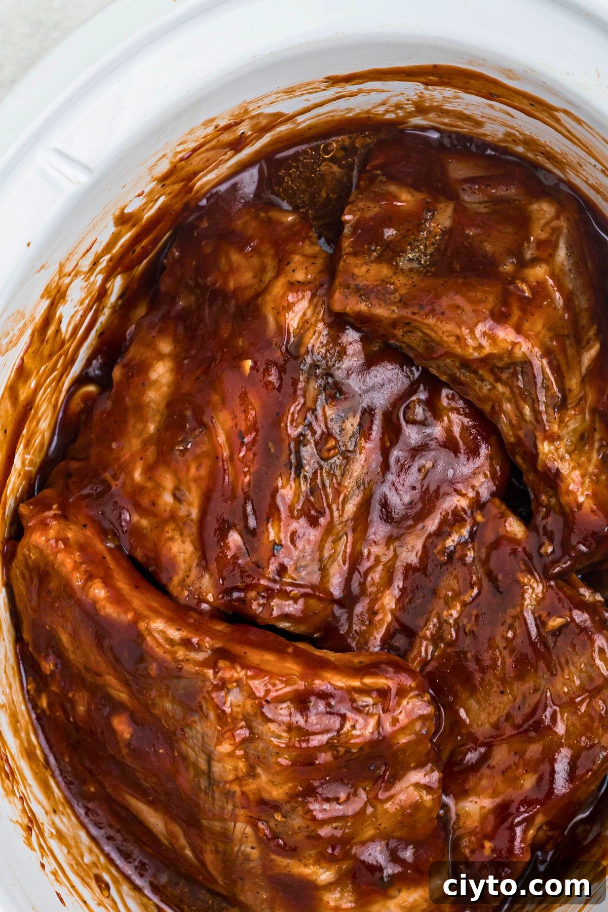 Juicy baby back ribs in a white slow cooker, tossed with BBQ sauce.