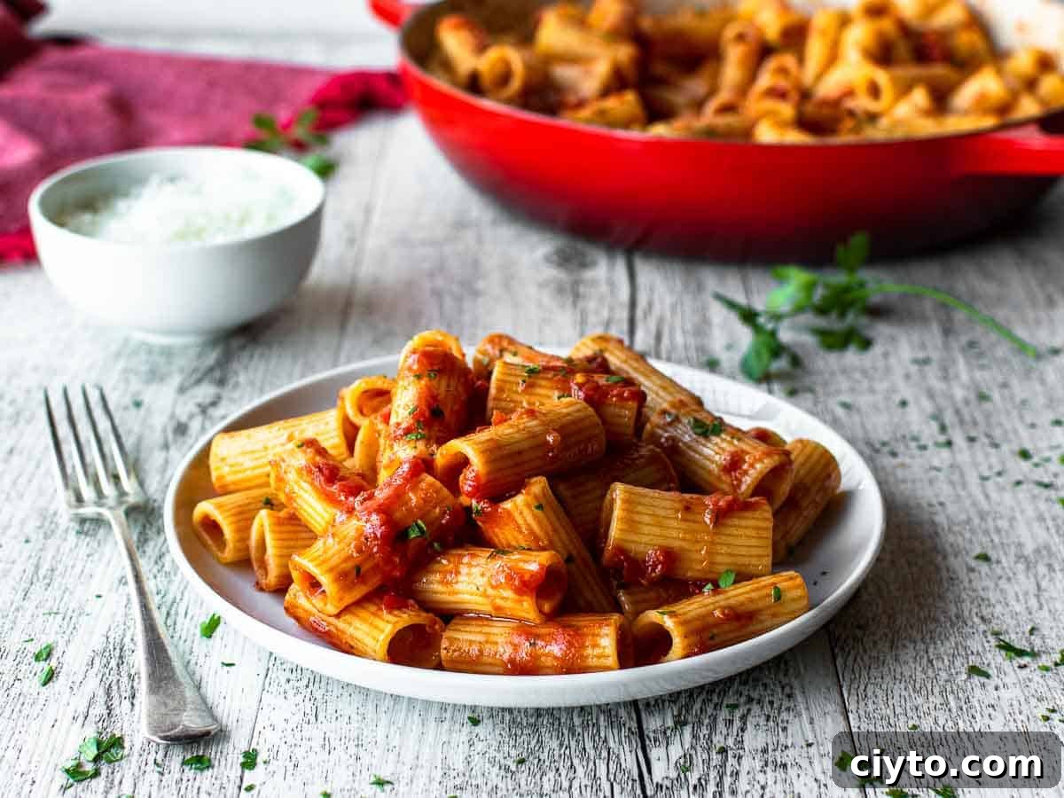 Rigatoni with tomato pasta sauce on a white plate with fork and red skillet in the background.