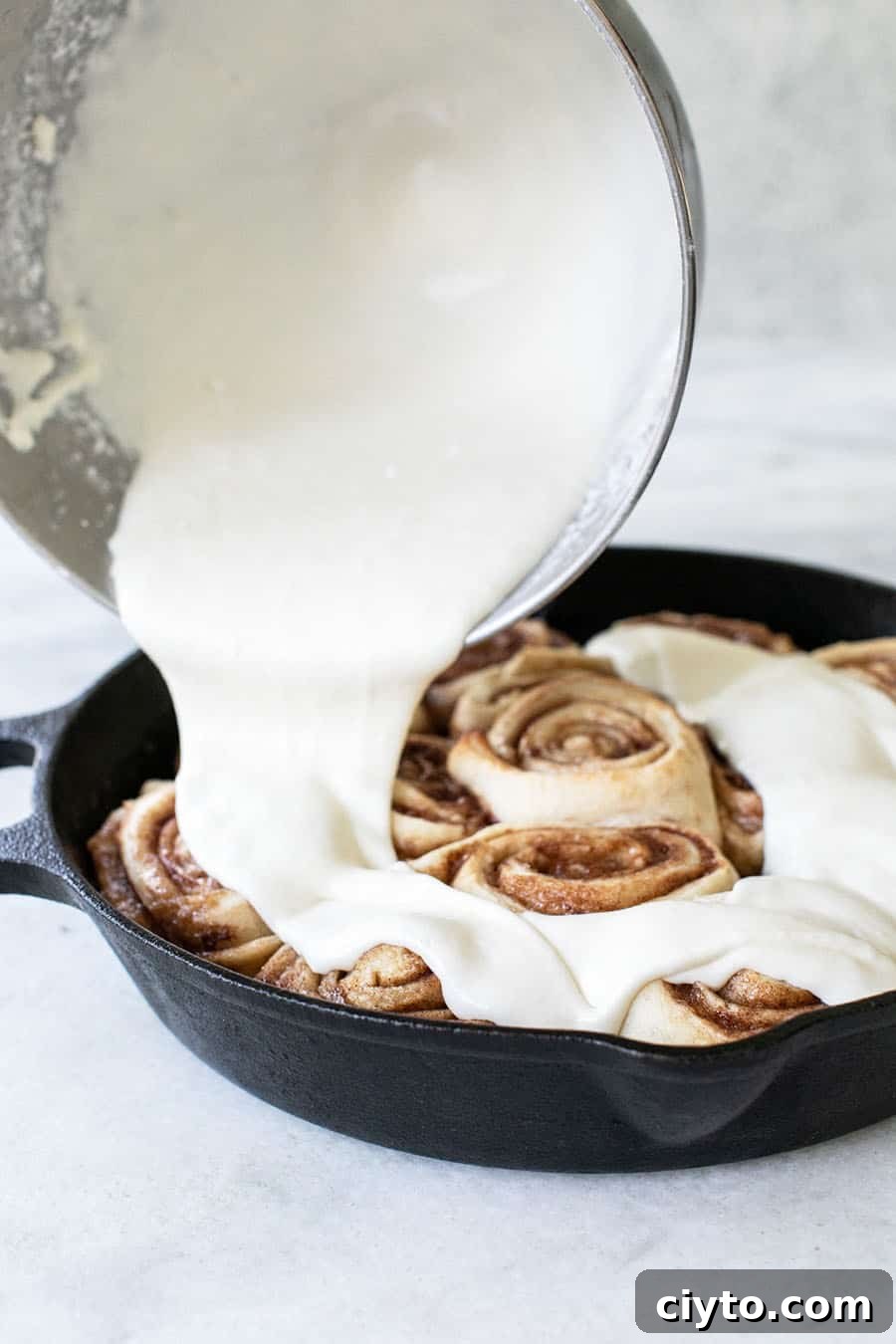 The glaze is poured over cinnamon rolls in a cast iron skillet.