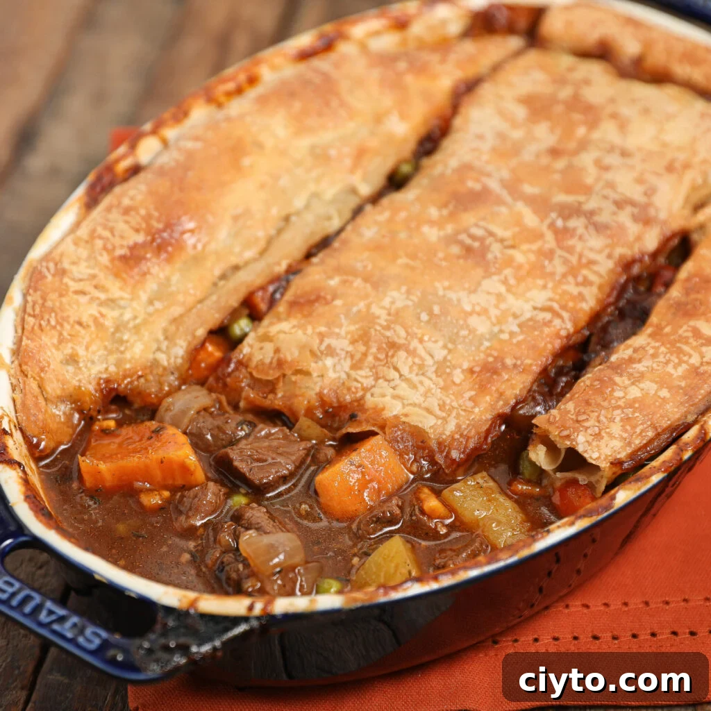 A hearty beef Guinness pot pie in a blue oval dish with a golden, flaky pastry crust, filled with chunks of beef, carrots, peas, and potatoes in rich brown gravy, set on a wooden surface with an orange napkin.