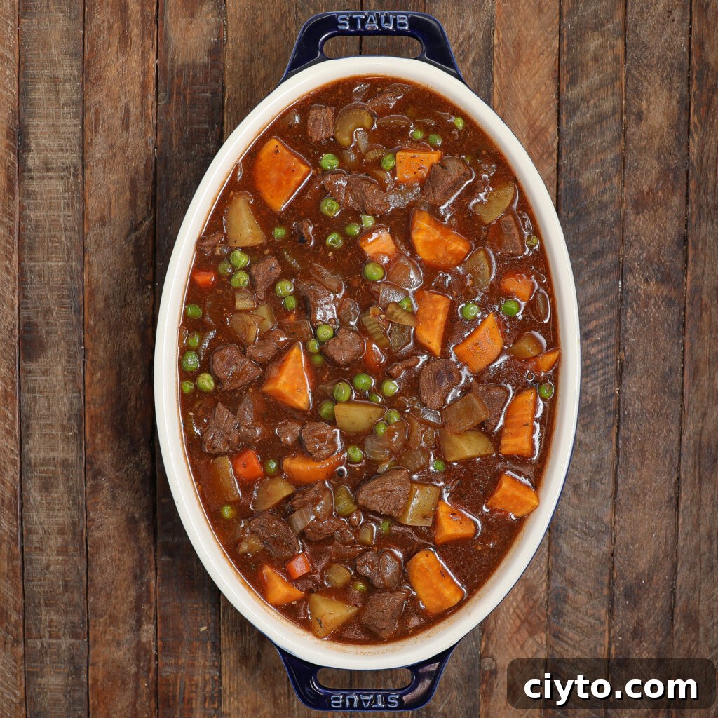 An oval dish filled with beef Guinness pot pie stew, featuring chunks of beef, potatoes, carrots, sweet potatoes, and green peas in a rich brown broth, set on a rustic wooden surface.