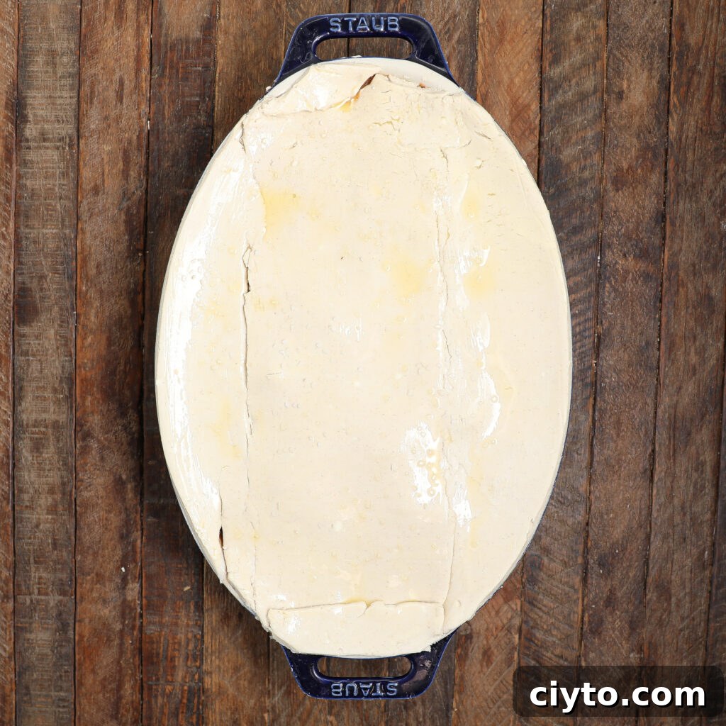 An oval blue baking dish filled with a beef Guinness pot pie, covered with an unbaked layer of pastry, sits on a wooden surface.