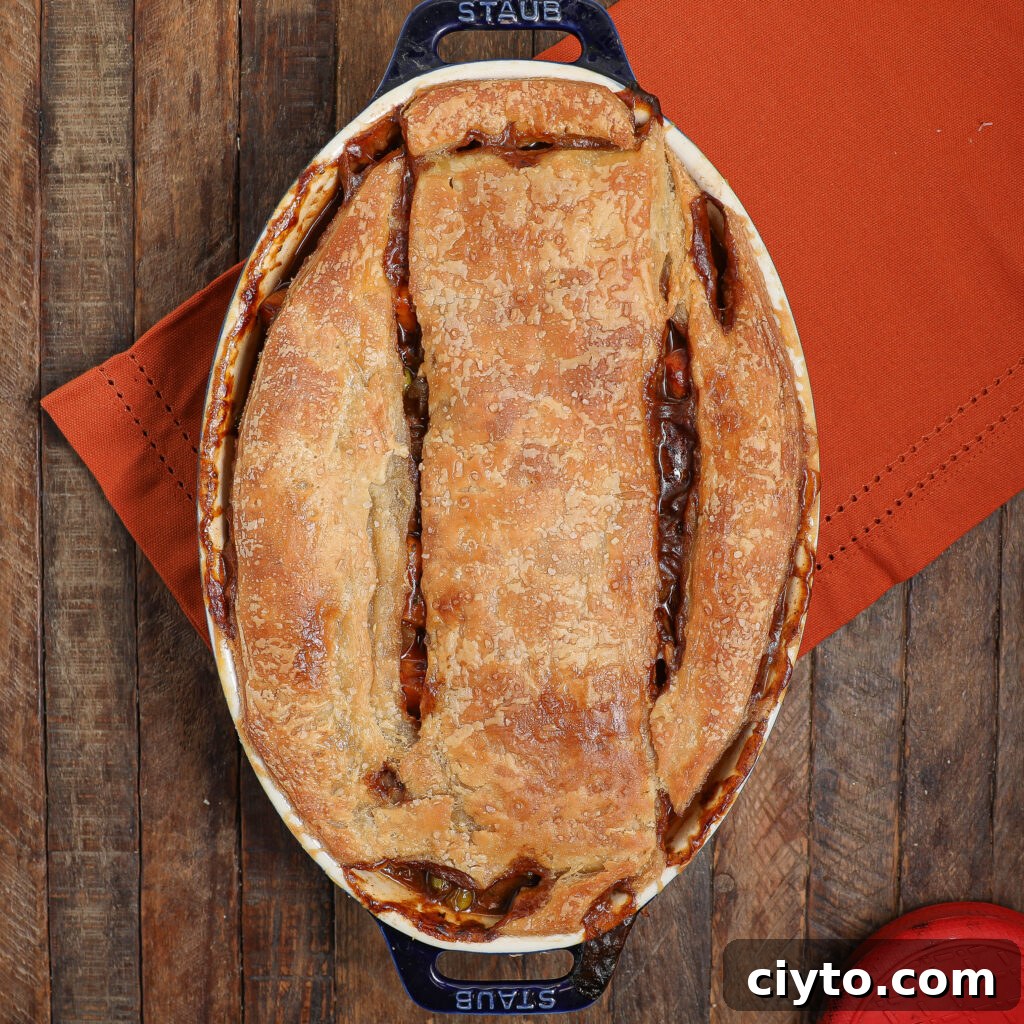 A golden-brown beef Guinness pot pie with a rustic crust sits in an oval dish on a wooden table, atop an orange cloth napkin. A red oven mitt is partially visible in the corner.