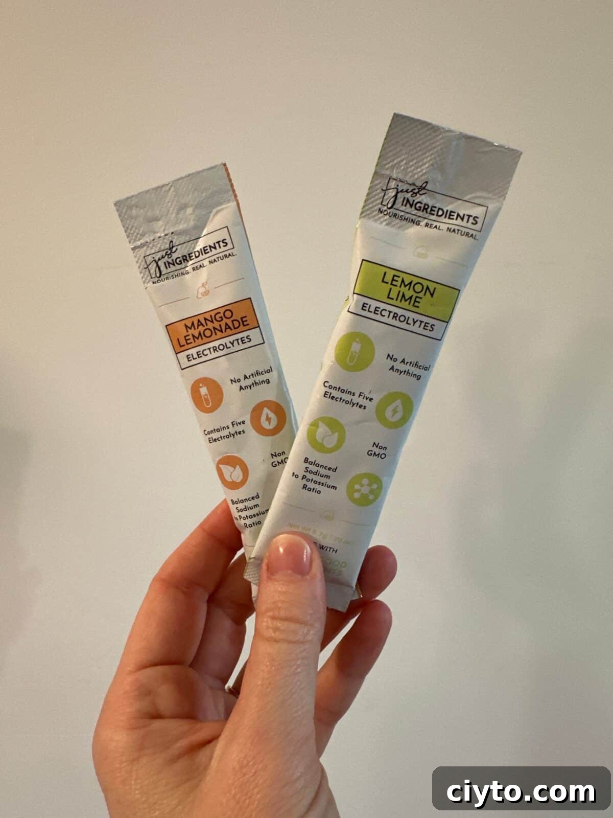 A person is holding two individual packets of "Just Ingredients" electrolyte drink mix, one in Mango Lemonade flavor (orange) and one in Lemon Lime flavor (green).