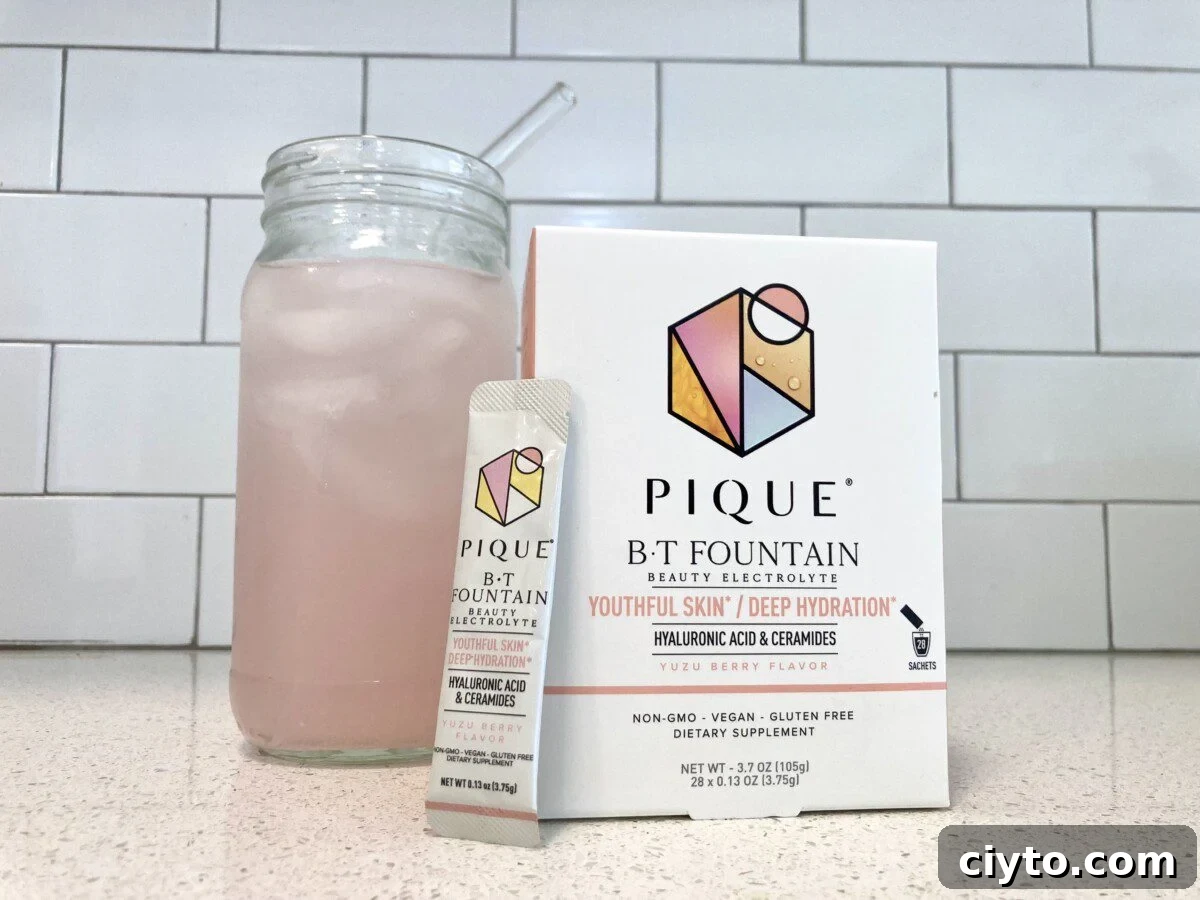 A glass of pink, prepared electrolyte drink sits next to a box and an individual sachet of Pique B-T Fountain Beauty Electrolyte in Yuzu Berry flavor.