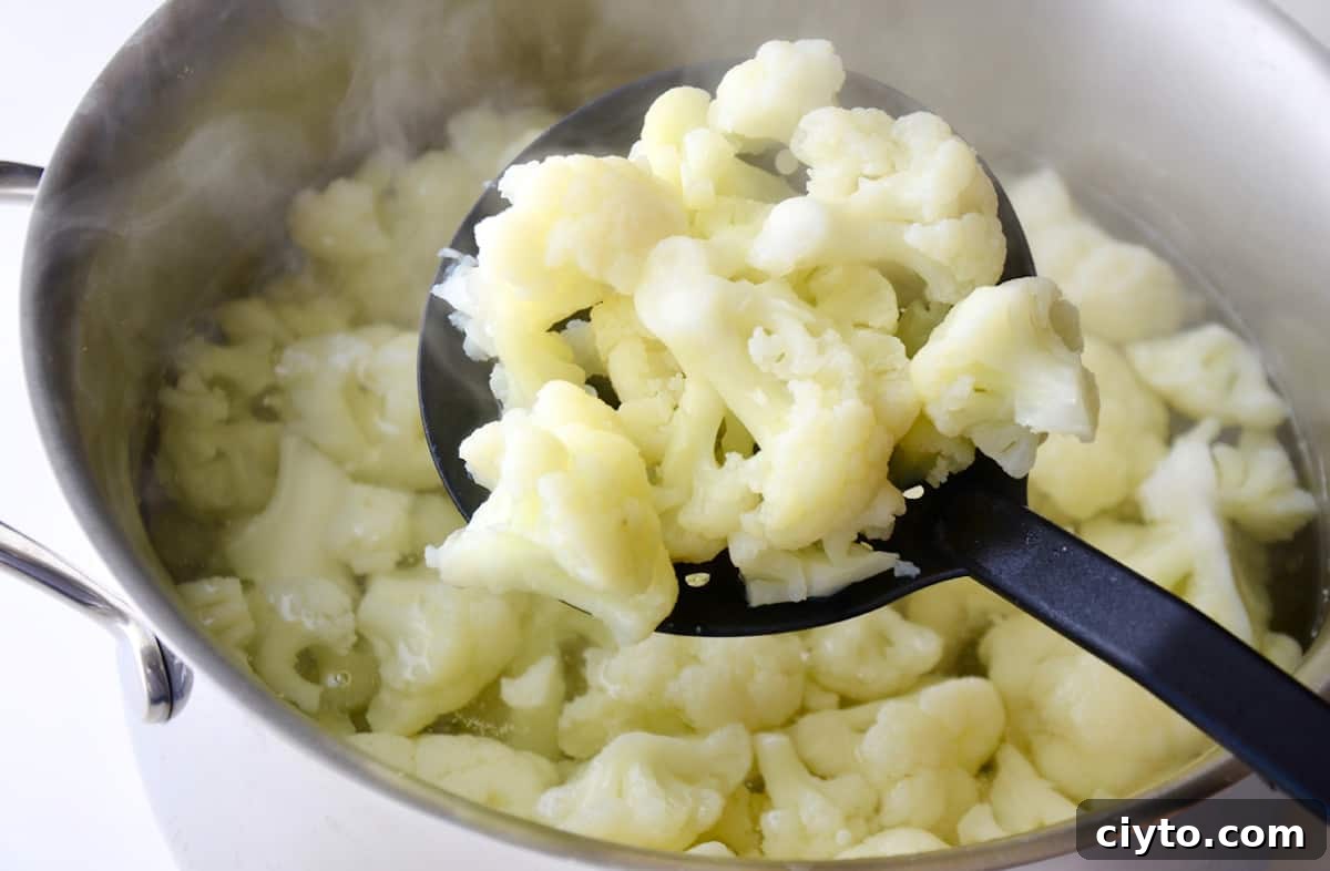 A slotted spoon holding cooked cauliflower florets over a large pot of boiling water containing more cooked cauliflower.