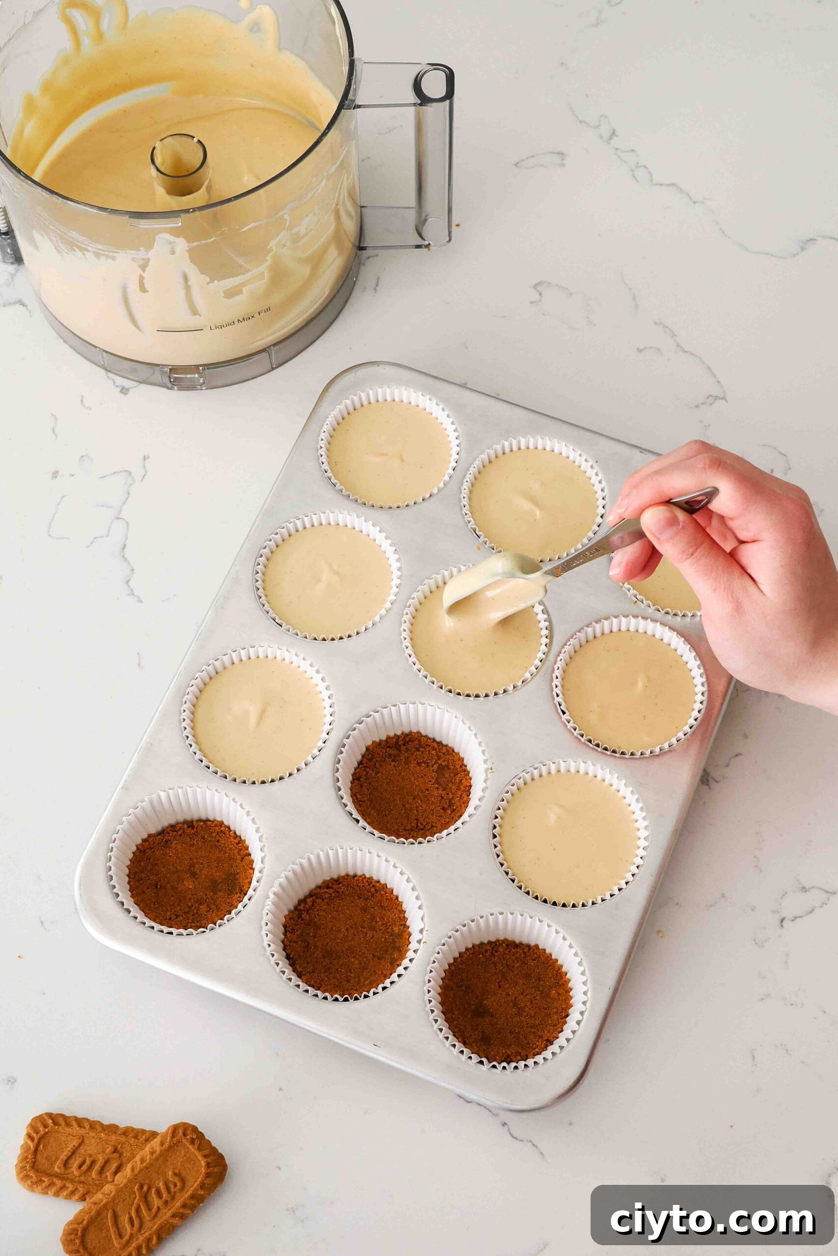 Mini Biscoff Cheesecakes Recipe with Cookie Butter Crust 12 A hand drops a Tablespoon of Biscoff cheesecake filling into paper liners in a muffin pan at a time.