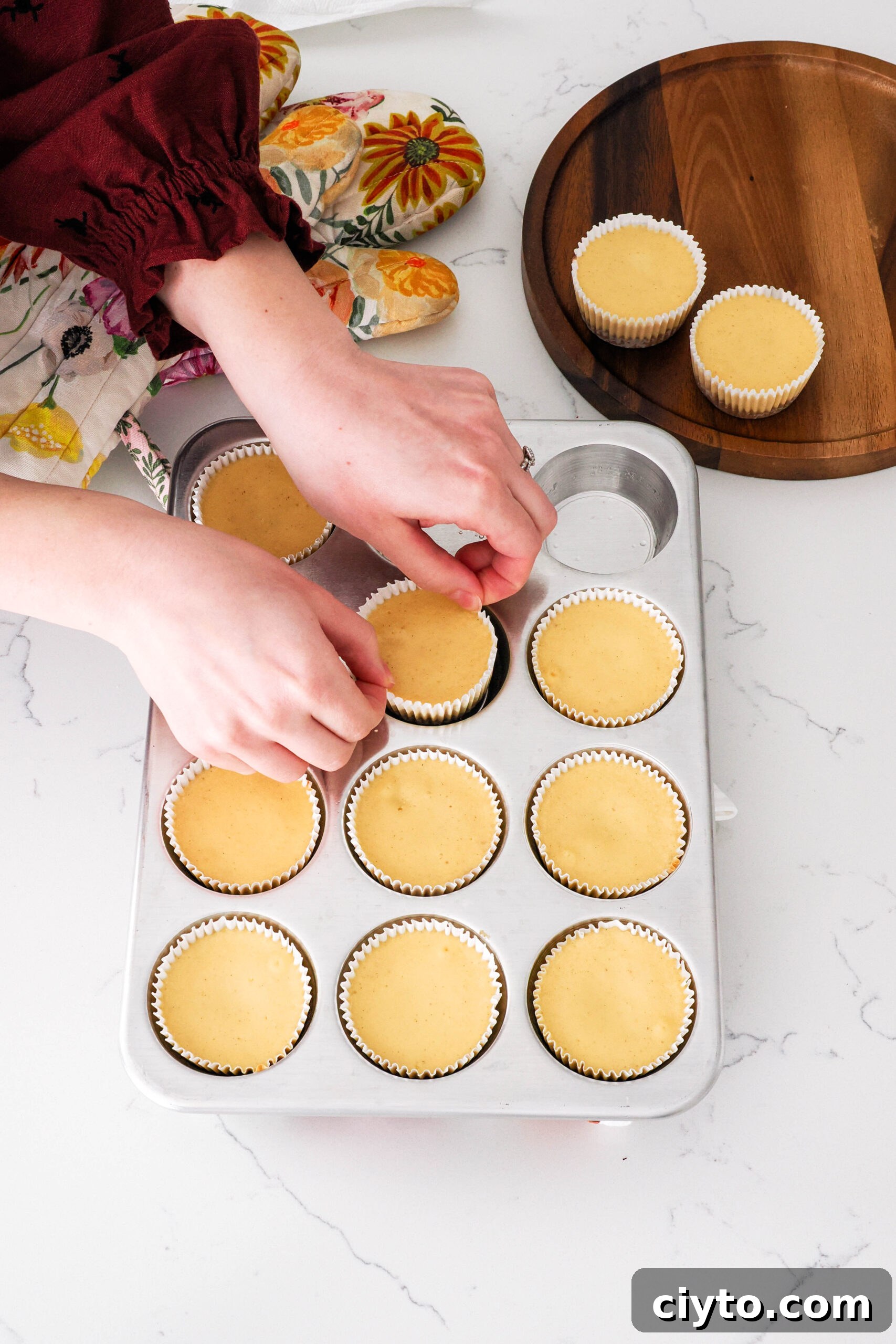 Mini Biscoff Cheesecakes Recipe with Cookie Butter Crust 15 Two hands pinch the edges of the paper liner on a mini cheesecake to remove it from a muffin pan.