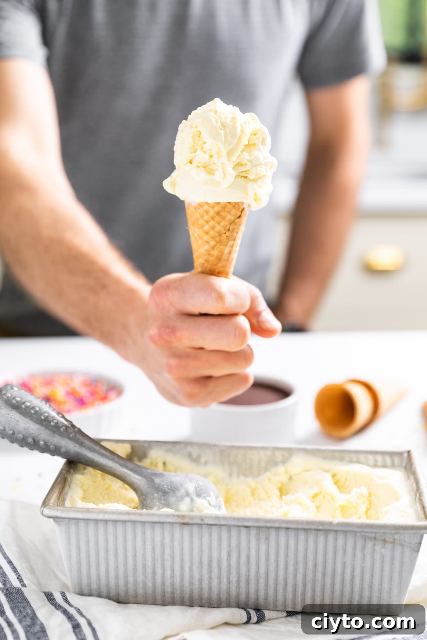 Creamy Vanilla Ice Cream Recipe with Real Vanilla Beans 2 Hand holding ice cream cone filled with vanilla ice cream with rest of vanilla ice cream in silver container below sitting on countertop