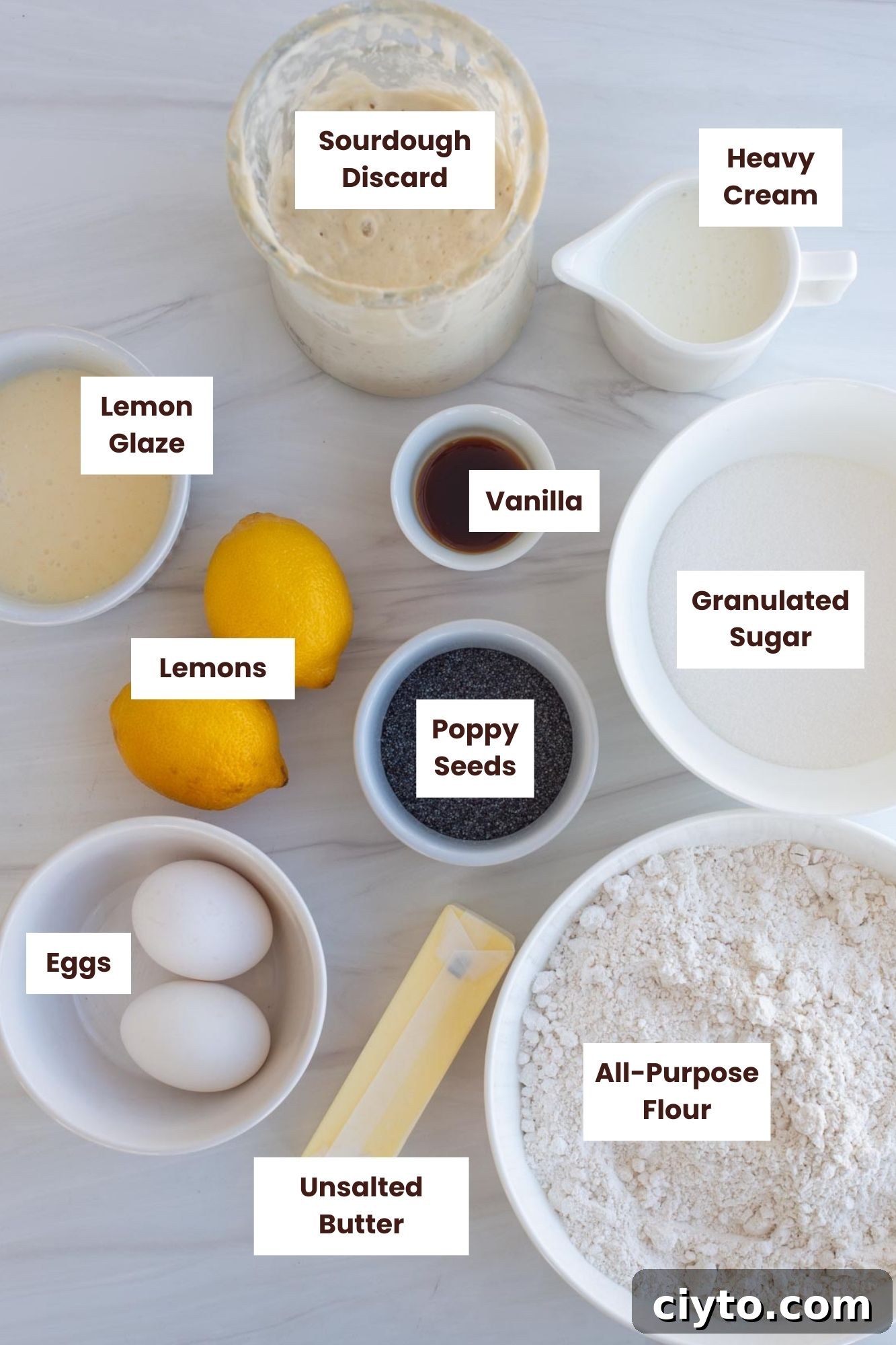 Lemon Poppy Seed Sourdough Scones Recipe 3 Ingredients are gathered on a countertop in separate containers, including sourdough discard, heavy cream, lemon glaze, vanilla, granulated sugar, poppy seeds, lemons, eggs, unsalted butter, and all-purpose flour.