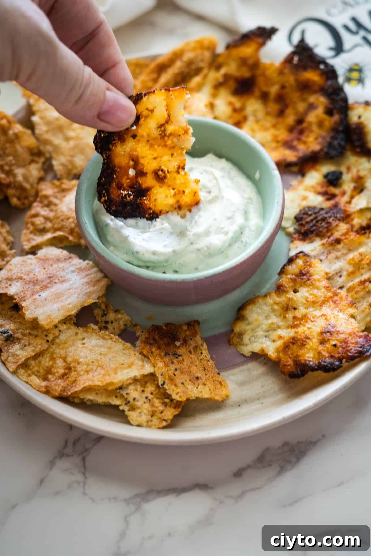 A hand dips a crispy snack into a bowl of creamy dip, surrounded by other similar snacks on a plate.
