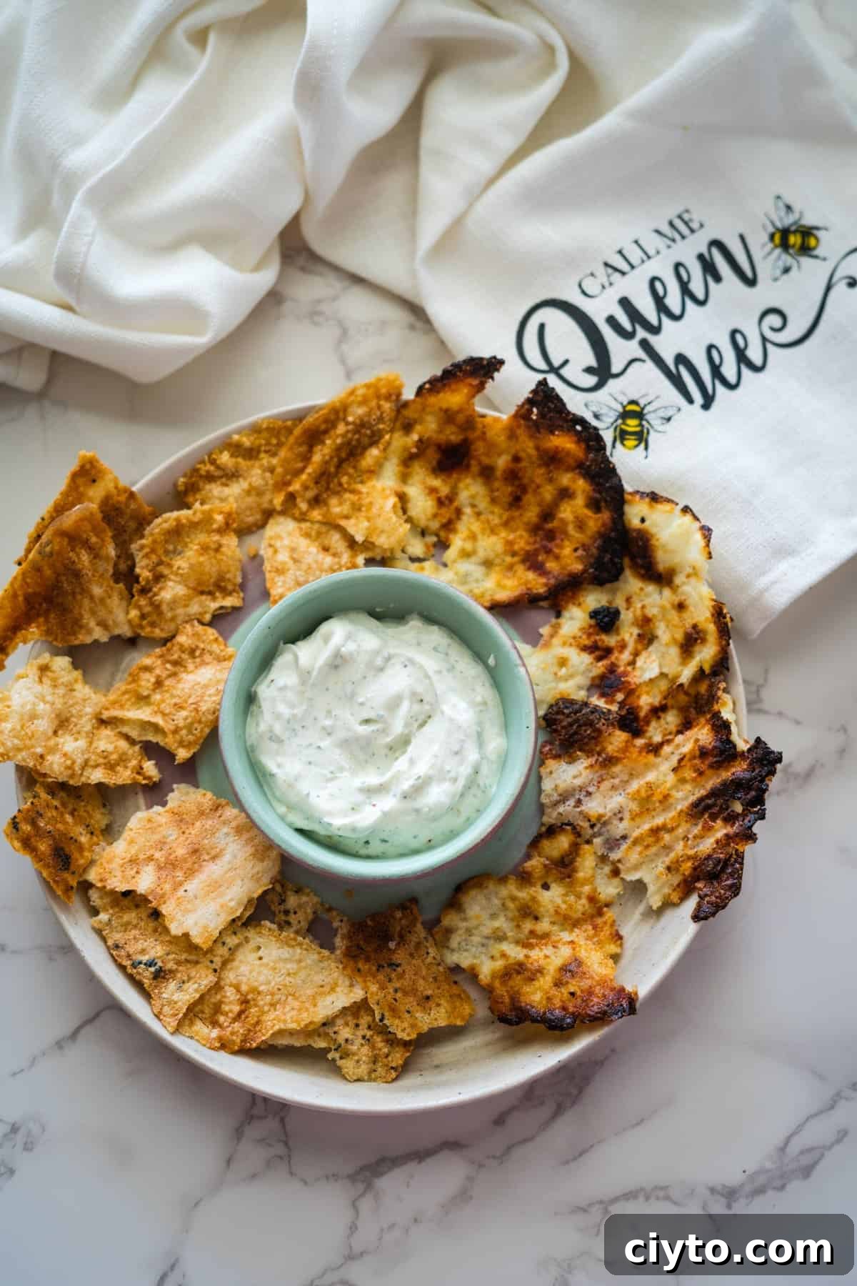 A plate of crispy bread slices surrounds a small bowl of white creamy dip, reminiscent of a luscious blend like cottage cheese. In the background, a napkin with the text "Call Me Queen Bee" and bee illustrations adds a whimsical touch.