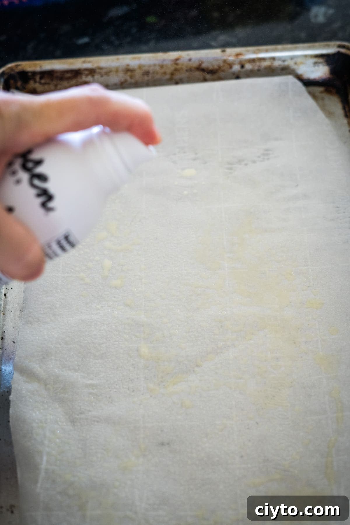 A person is spraying a can of oil onto a baking tray lined with parchment paper.
