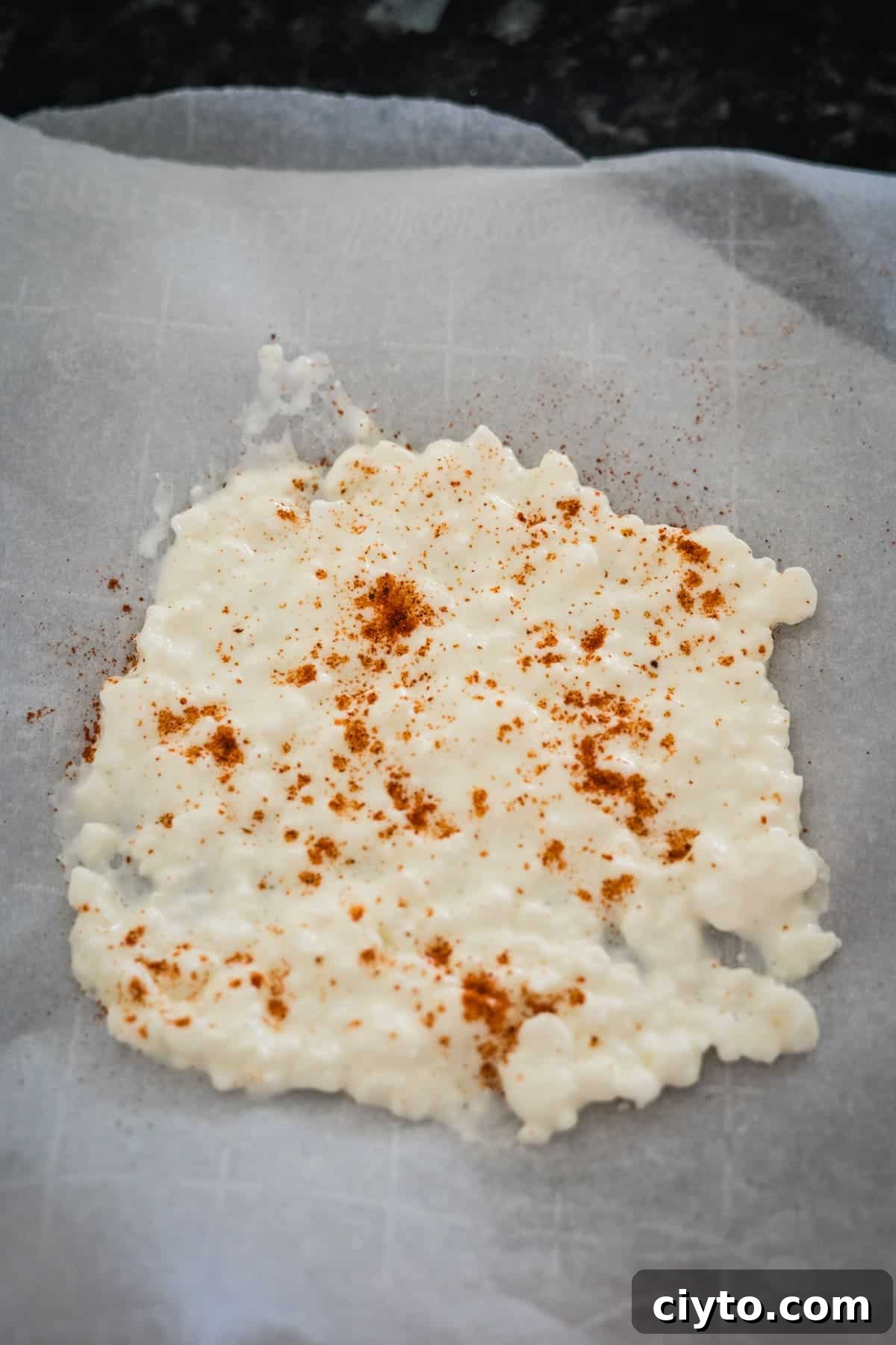 Parchment paper with white melted cheese sprinkled with a small amount of paprika, on a dark countertop.