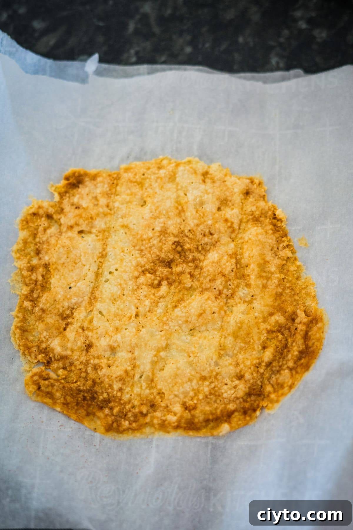 A baked, golden-brown flatbread or crust on a piece of parchment paper resting on a dark surface.