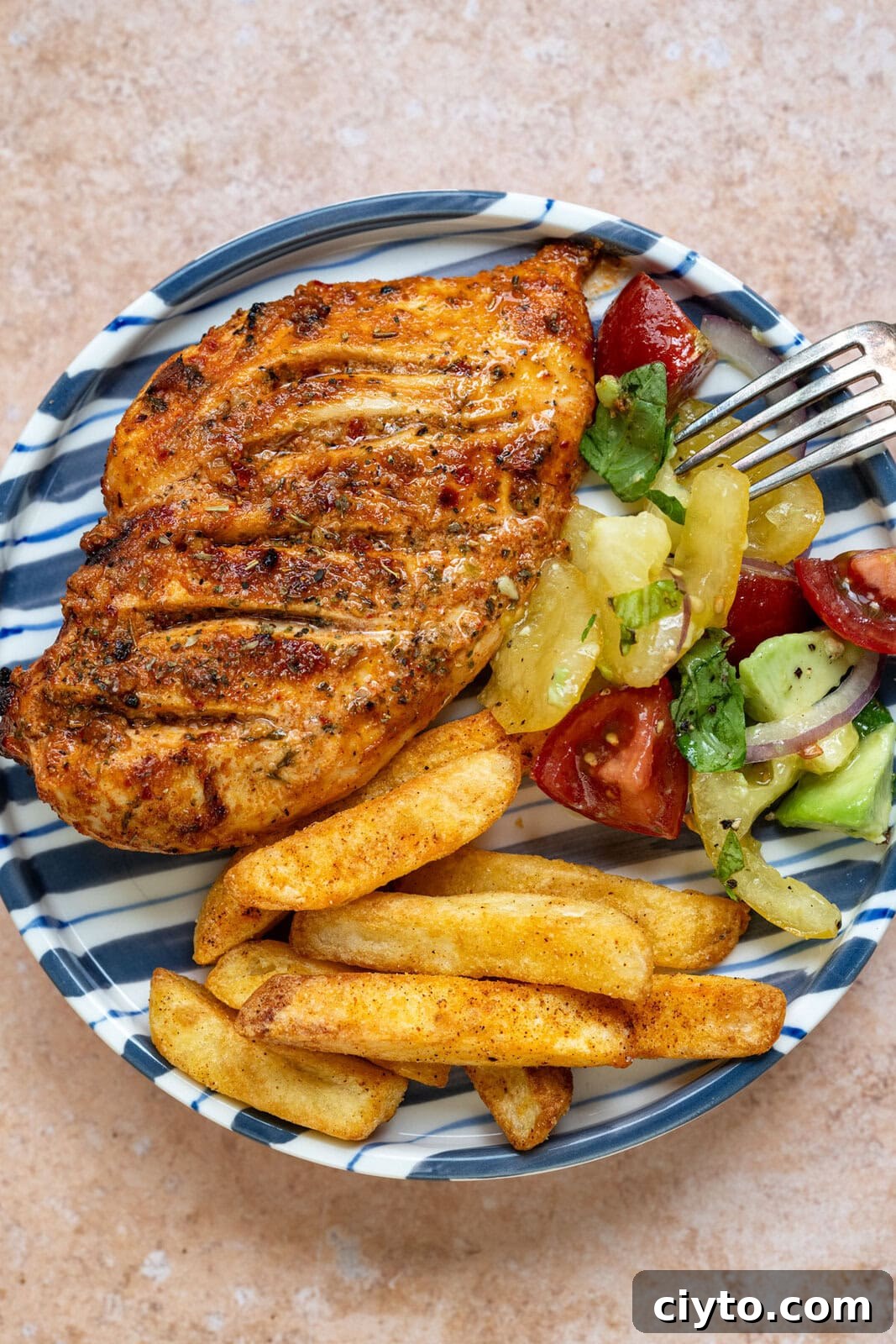 How to Make Juicy Air Fryer Chicken Breast 3 A plate with air fryer chicken breast, seasoned potato fries, and a fresh salad of tomatoes, cucumbers, and greens. A fork rests on the plate. The plate has a blue-and-white striped rim on a beige surface.