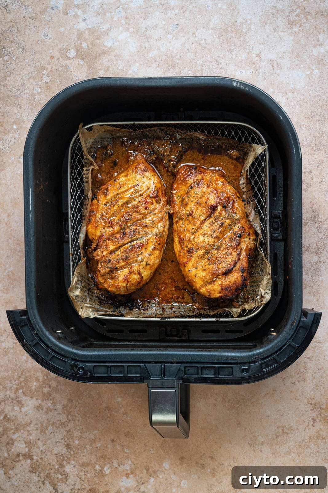How to Make Juicy Air Fryer Chicken Breast 7 Two seasoned, cooked chicken breasts in an air fryer basket lined with parchment paper, resting on a light brown countertop.