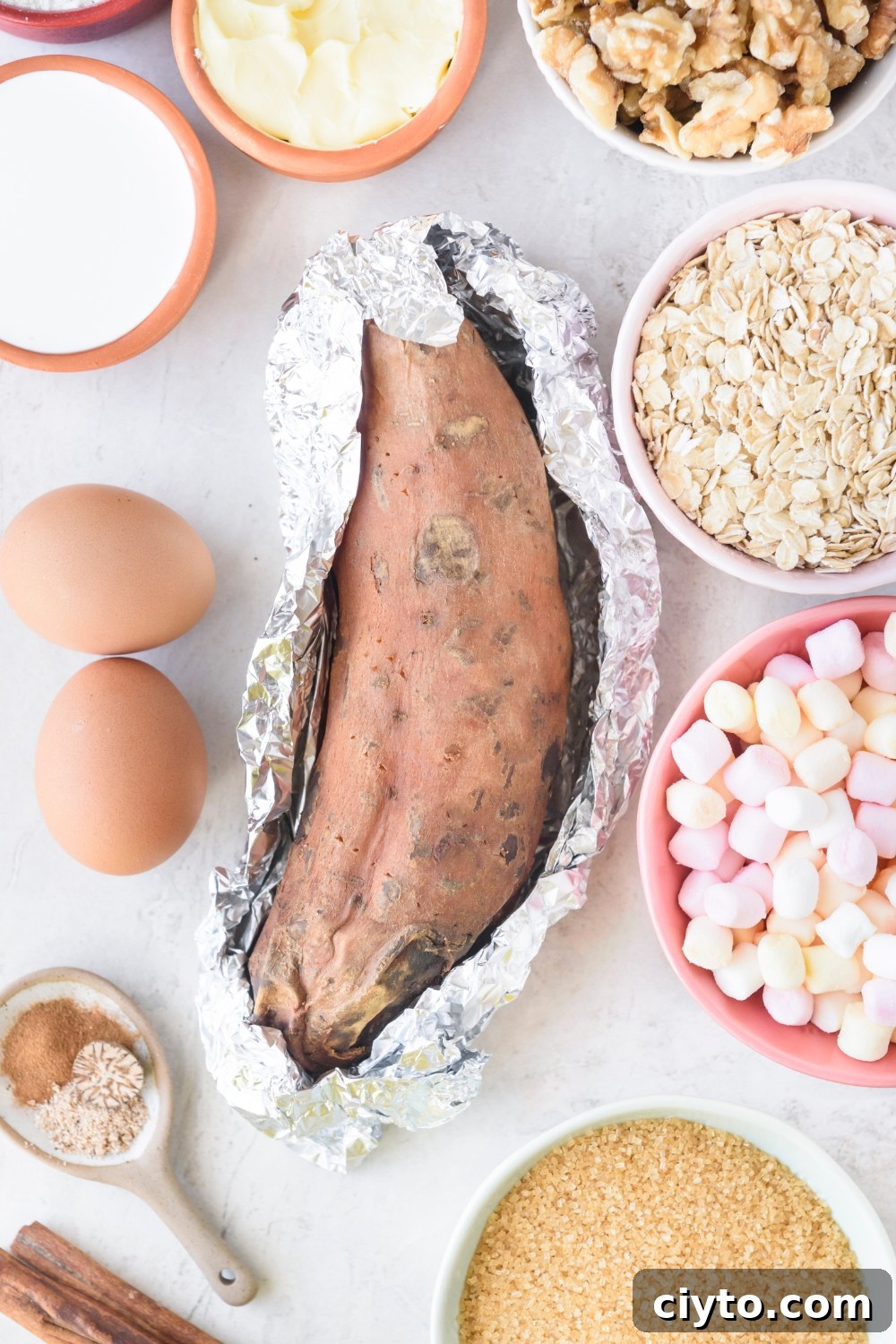 Copycat Boston Market Sweet Potato Casserole Recipe 5 One large sweet potato partially wrapped in foil is on a white counter. Surrounding the potato are bowls of walnuts, oats, marshmallows, brown sugar, flour, cream, and two eggs.