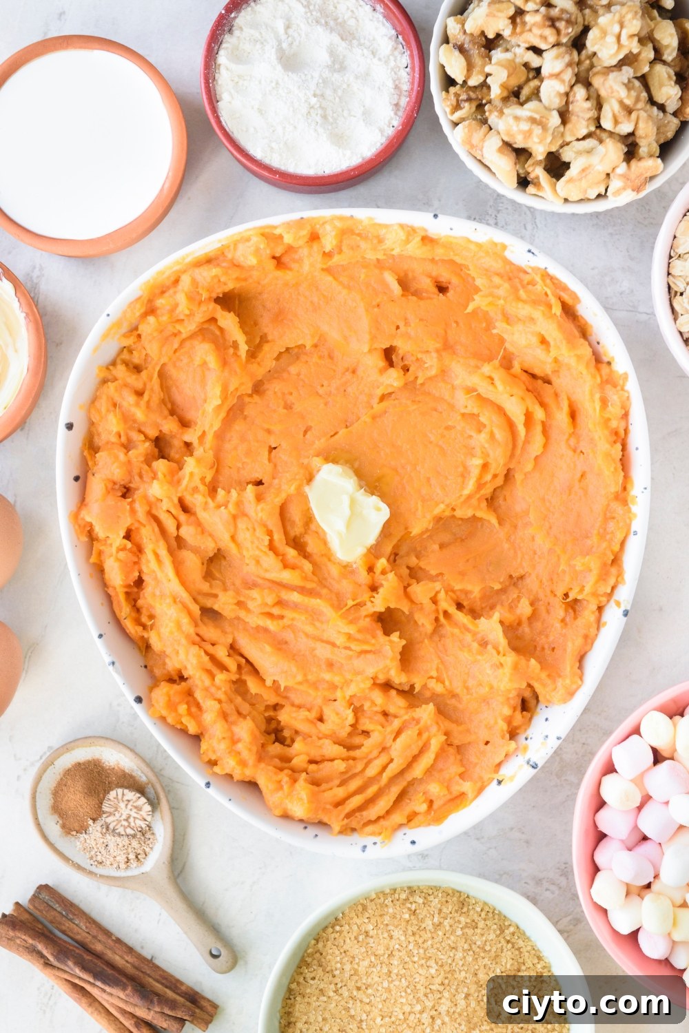 Copycat Boston Market Sweet Potato Casserole Recipe 6 A large white bowl full of mashed sweet potatoes with a pat of butter in the middle. Bowls full of walnuts, marshmallows, brown sugar, spices, flour, and cream surround the large bowl