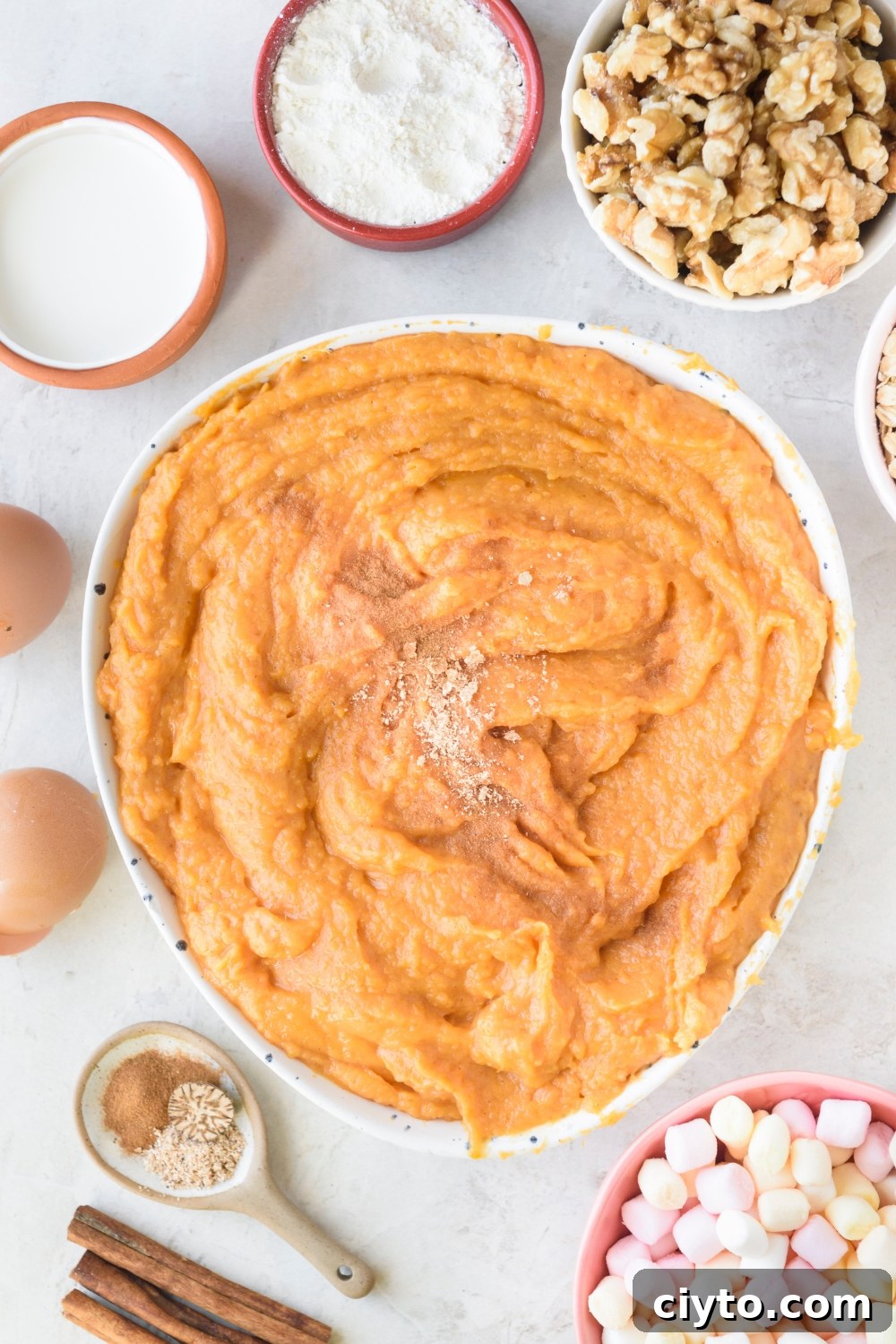 Copycat Boston Market Sweet Potato Casserole Recipe 7 A large bowl of mashed sweet potatoes with spices on top on a white counter. Bowls of walnuts, flour, cream, 2 eggs, spices, and mini marshmallows surround the bowl.