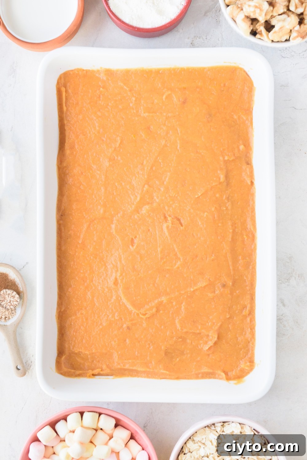 Copycat Boston Market Sweet Potato Casserole Recipe 8 A white casserole dish is full of mashed sweet potatoes in an even layer. Bowls of marshmallows, oats, walnuts, flour, and cream surround the dish.