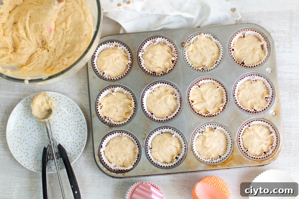 a cookie scoop sits beside a bowl of cupcake batter and 12, filled cupcakes liners in a tray ready for the oven