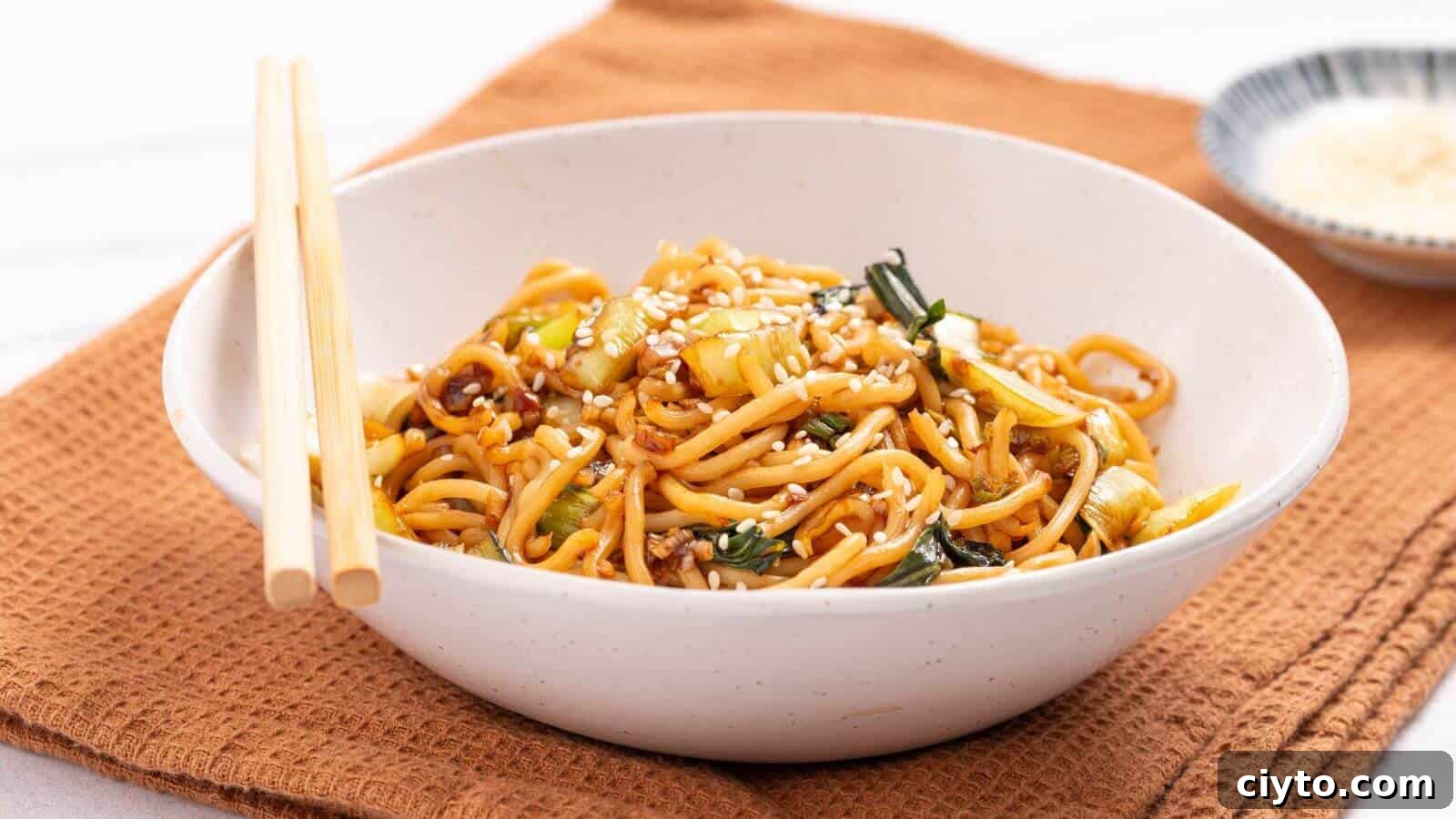 A bowl of stir-fried noodles with vegetables, topped with sesame seeds, placed on a brown cloth napkin. Chopsticks rest on the bowl's edge, and a small dish is visible in the background.