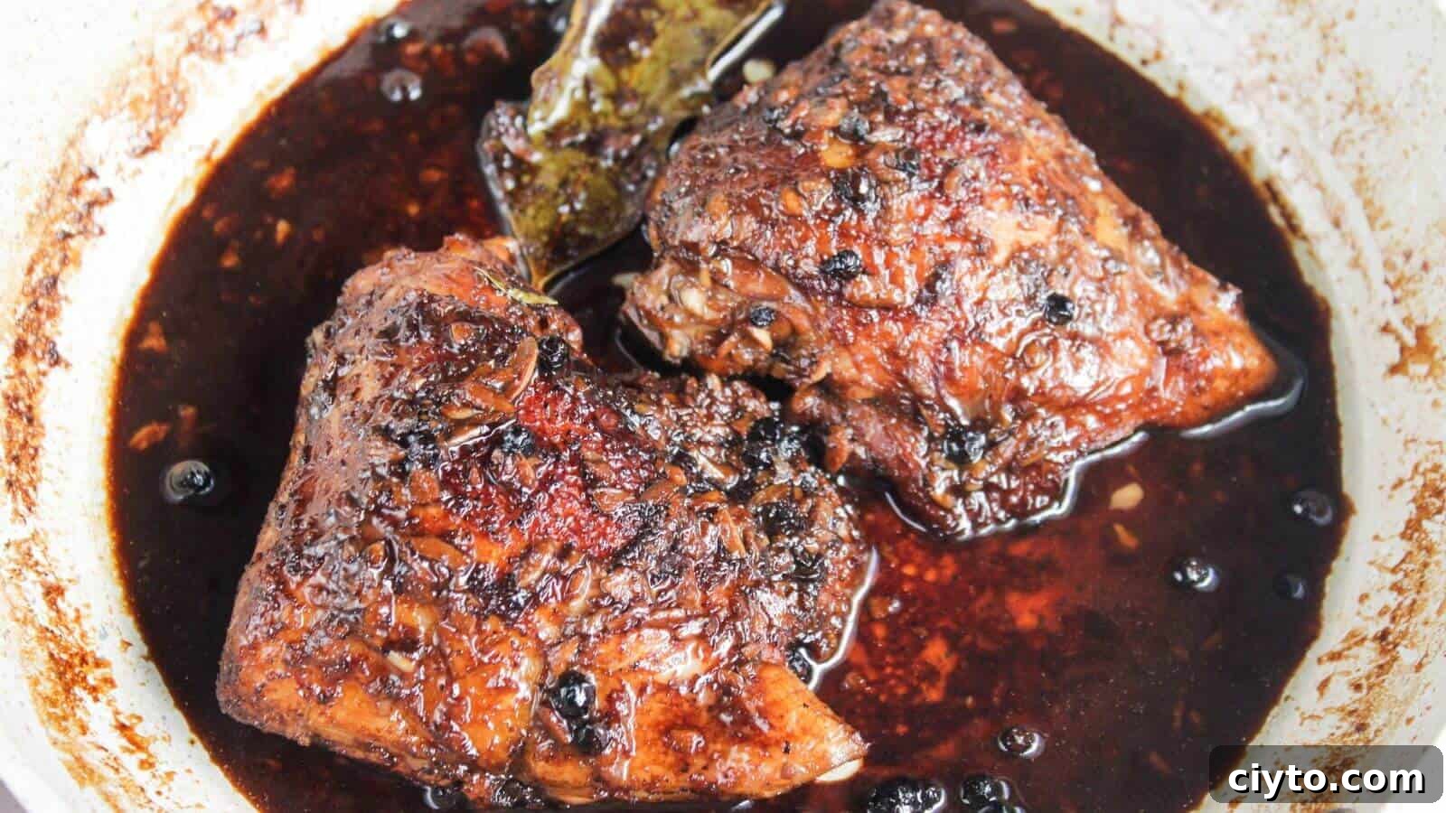 Two pieces of braised meat in a dark, rich sauce, seasoned with whole peppercorns and a bay leaf, in a pan.