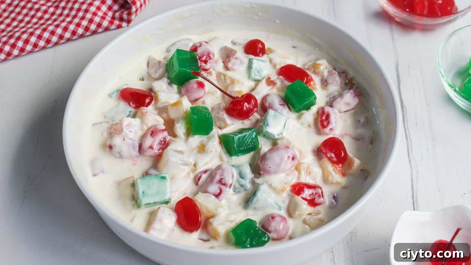 A white bowl filled with fruit salad in creamy sauce, topped with green gelatin cubes and maraschino cherries, sits on a white surface.