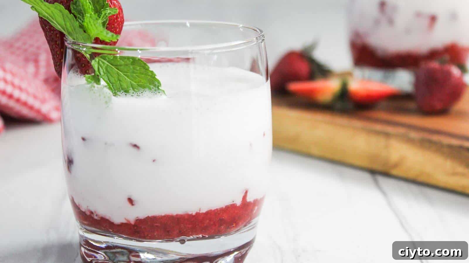 A glass of strawberry milk garnished with a fresh strawberry and mint leaf, set on a white surface.