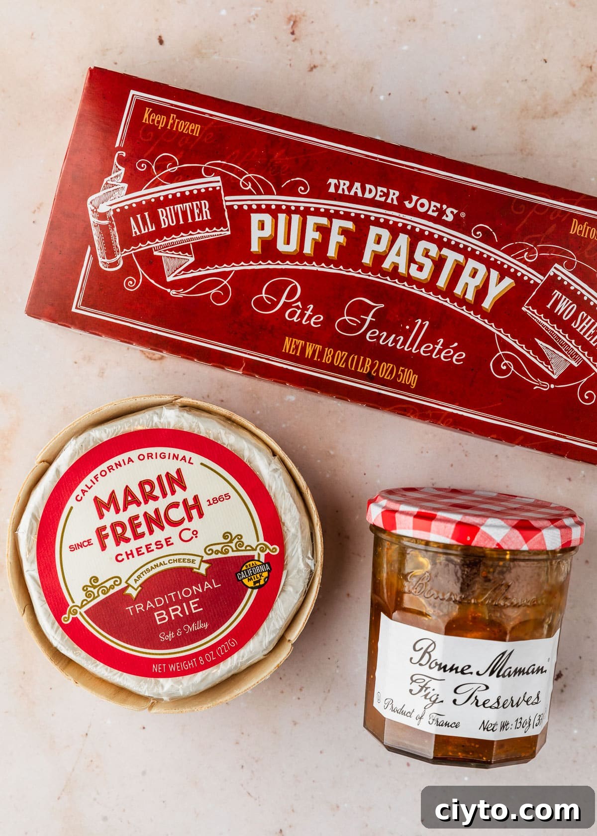 3-Ingredient Brie and Jam Puff Pastry Bites 3 A box of puff pastry, a wheel of brie cheese, and a jar of preserves on a beige marble counter.