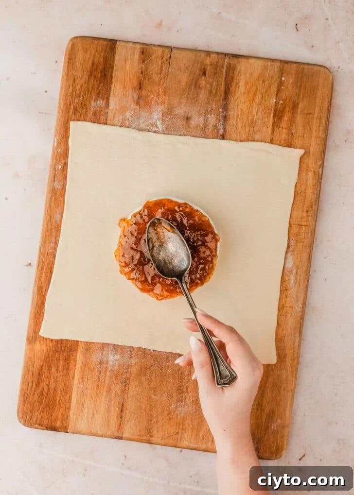 3-Ingredient Brie and Jam Puff Pastry Bites 6 A hand using a spoon to spread fig preserves on cheese in the middle of a square of pastry.