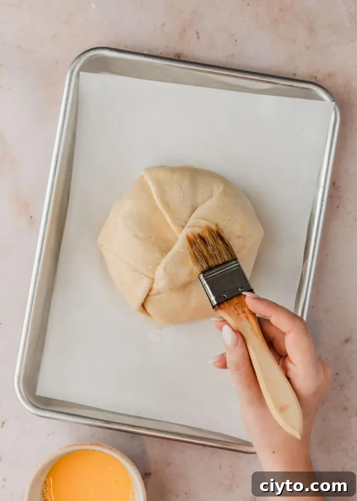 3-Ingredient Brie and Jam Puff Pastry Bites 8 A hand using a pastry brush to brush egg wash on brie en croute on a sheet pan.