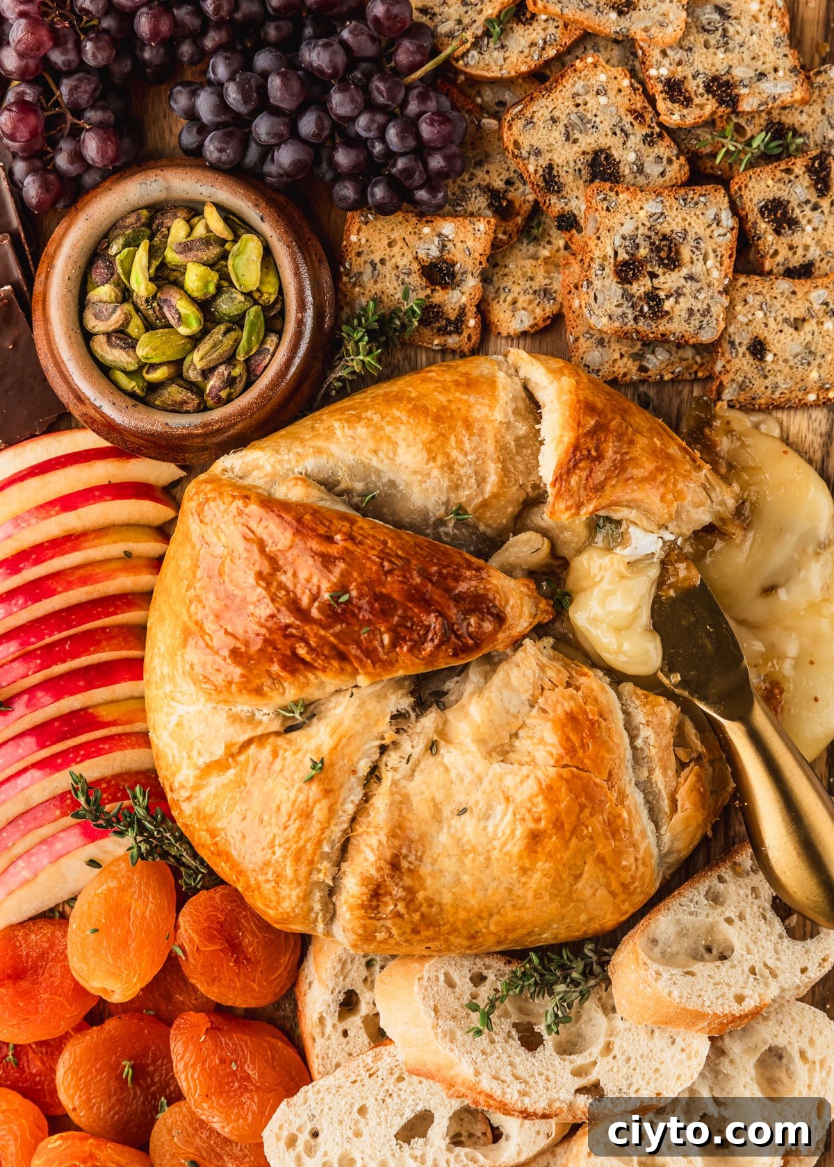 3-Ingredient Brie and Jam Puff Pastry Bites 10 Brie and jam in puff pastry on a wood board next to crackers, apples, grapes, dried apricots, and a brown bowl of pistachios.